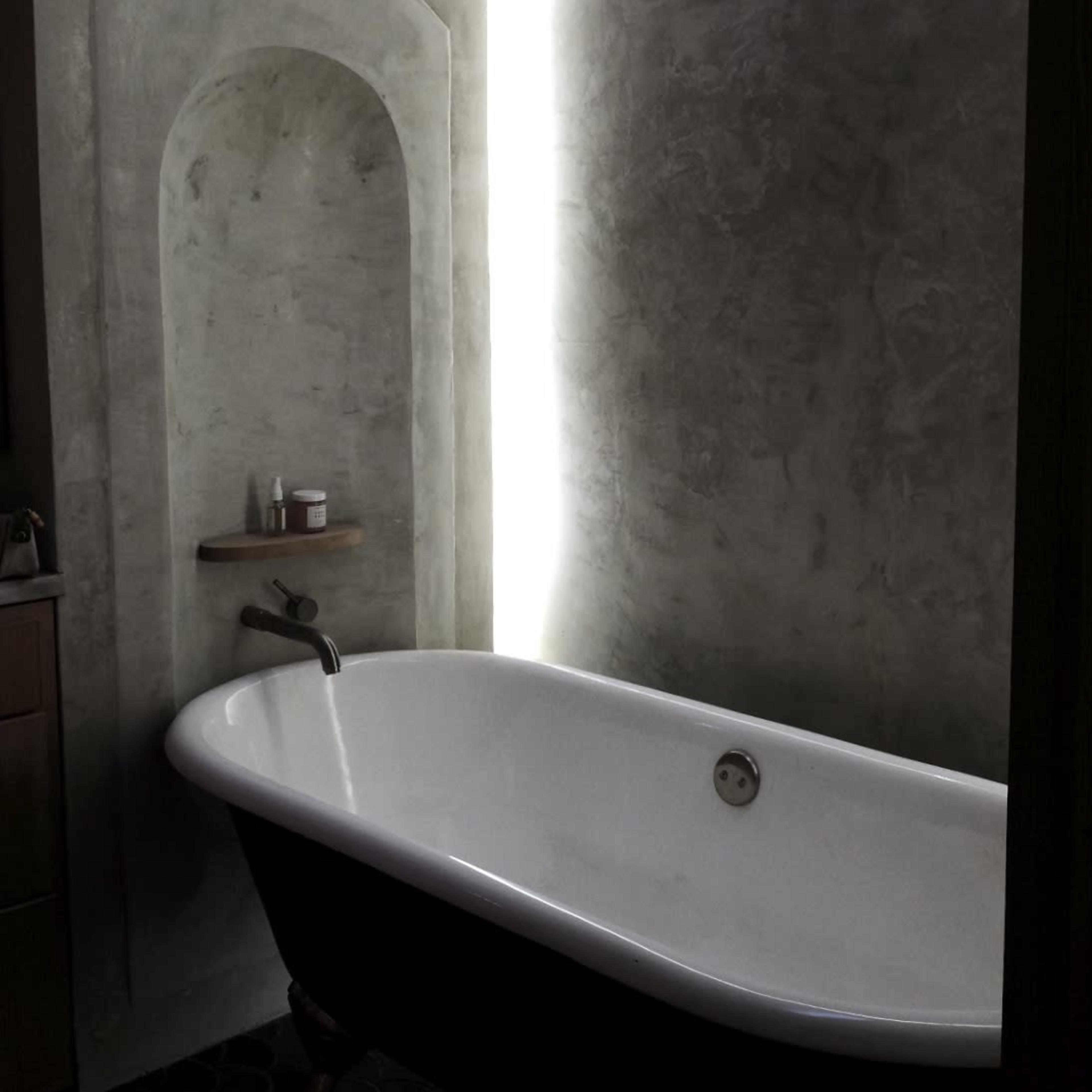 A freestanding bathtub is positioned beside a curved, illuminated wall in a modern bathroom with a concrete finish.