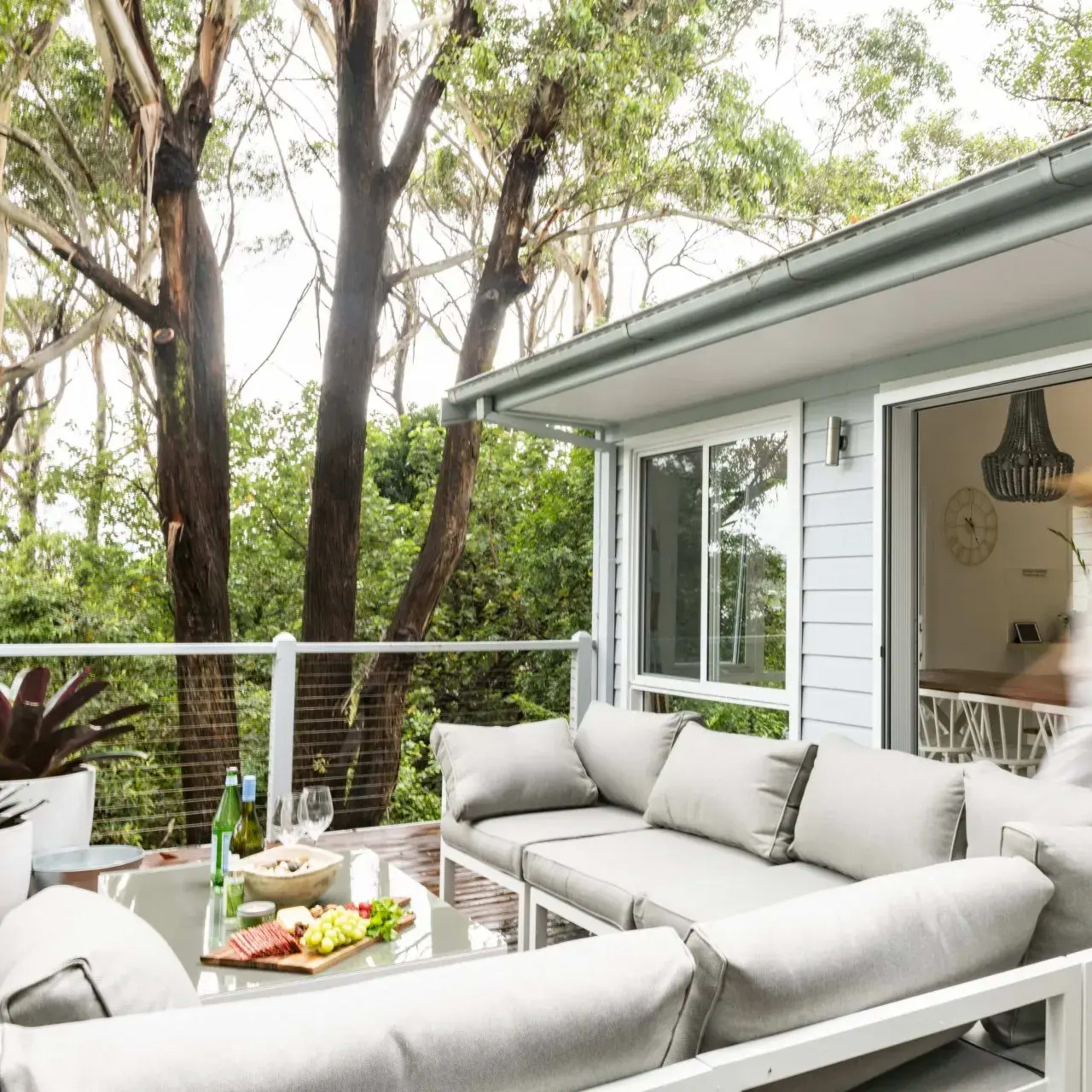 A person walks through an open doorway from a deck area with a large gray sofa into a kitchen and dining space surrounded by trees.