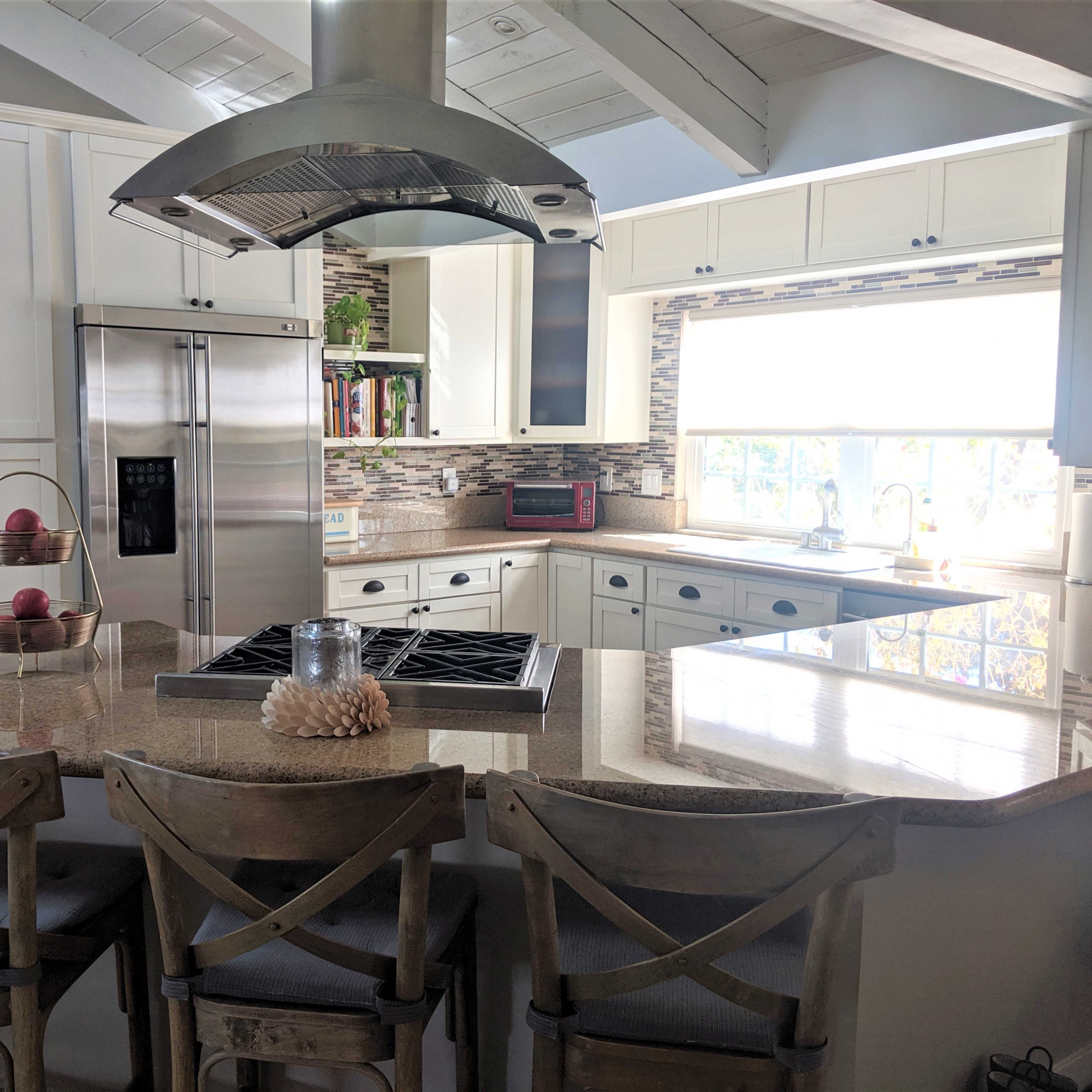 A modern kitchen with a central island, featuring a gas stovetop, three wooden stools, and white cabinetry with a window that highlights the countertop and backsplash.