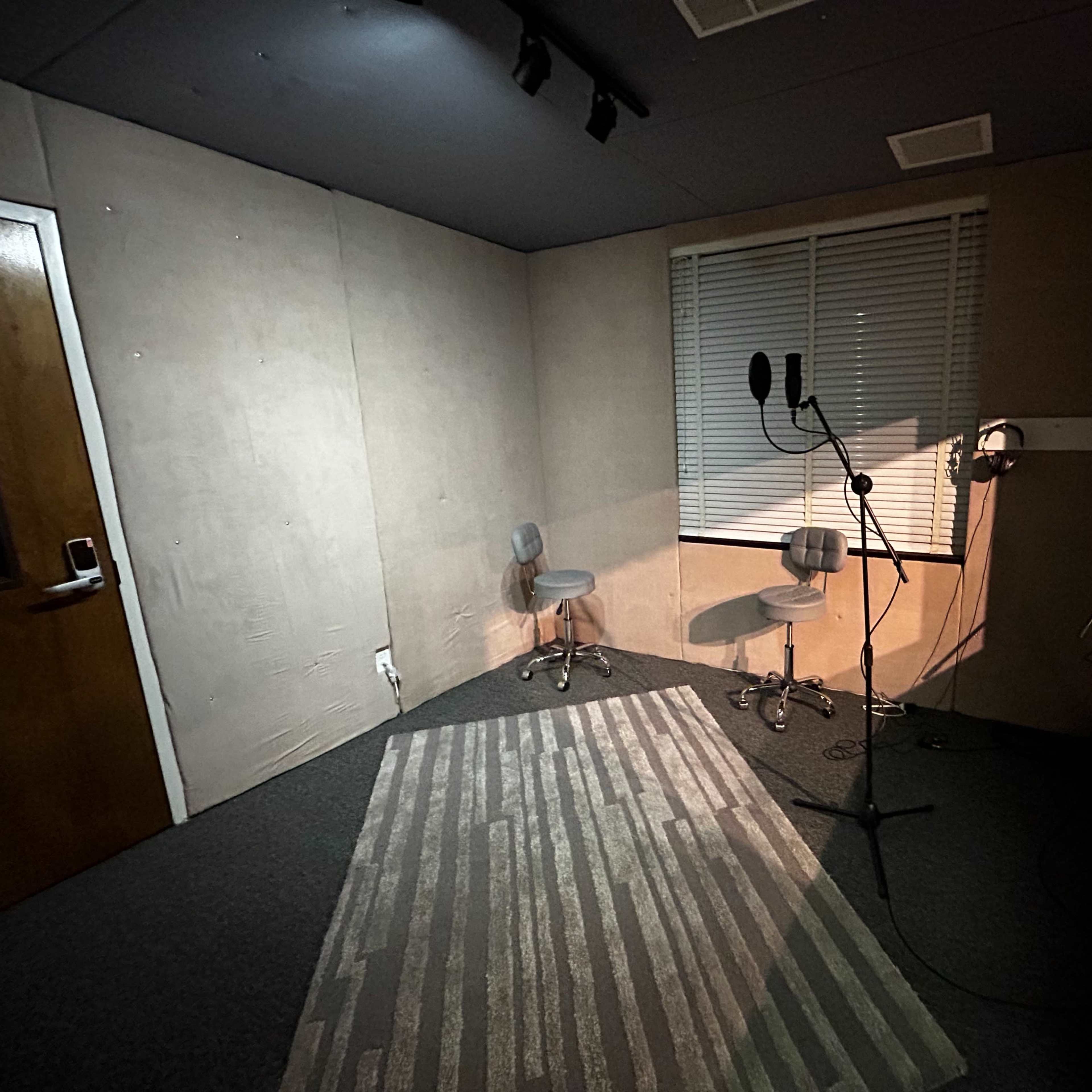 A small recording studio with two chairs, a microphone setup, and a window overlooking another room.