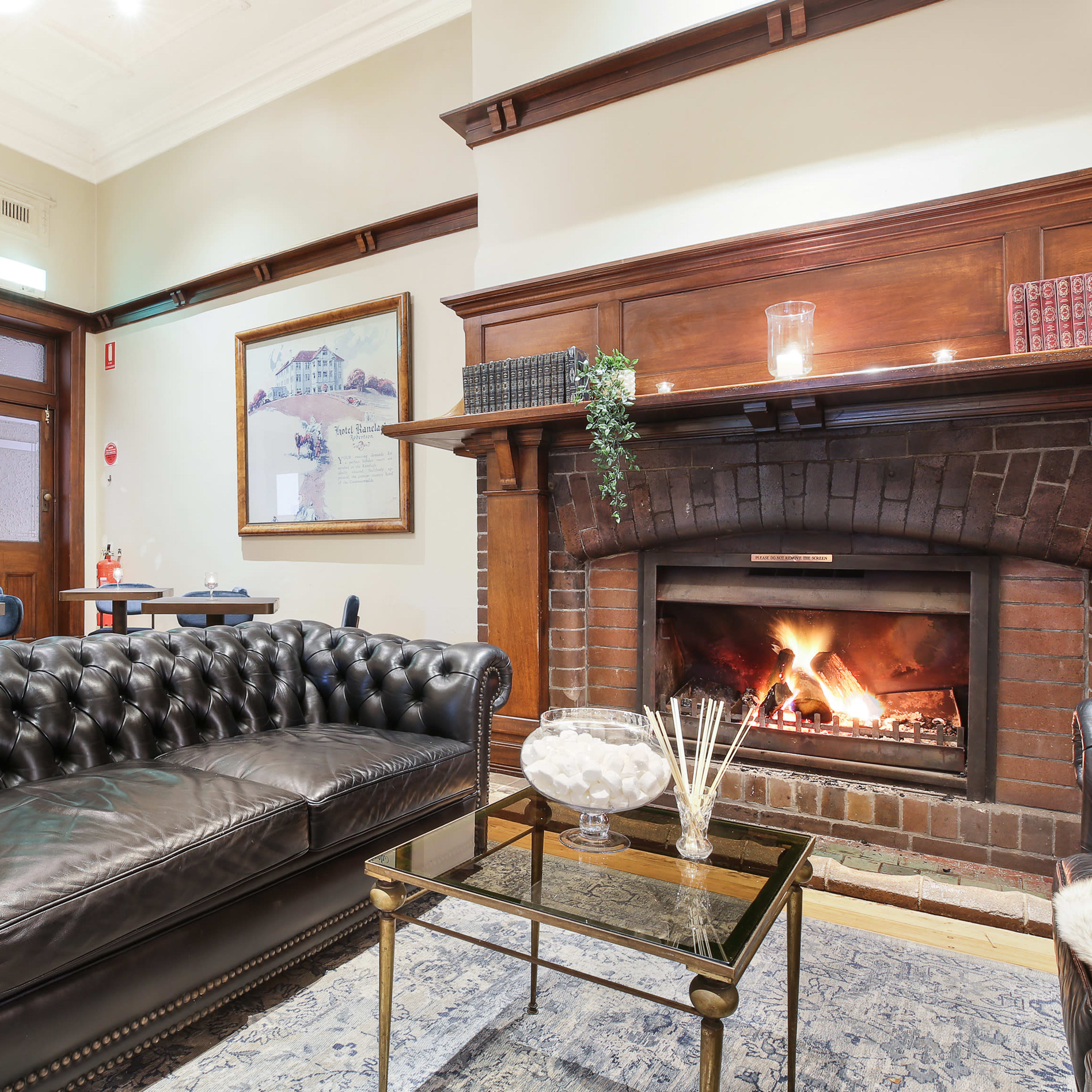 A cozy lounge area featuring a leather chesterfield sofa, a glass coffee table, and a brick fireplace with flames.