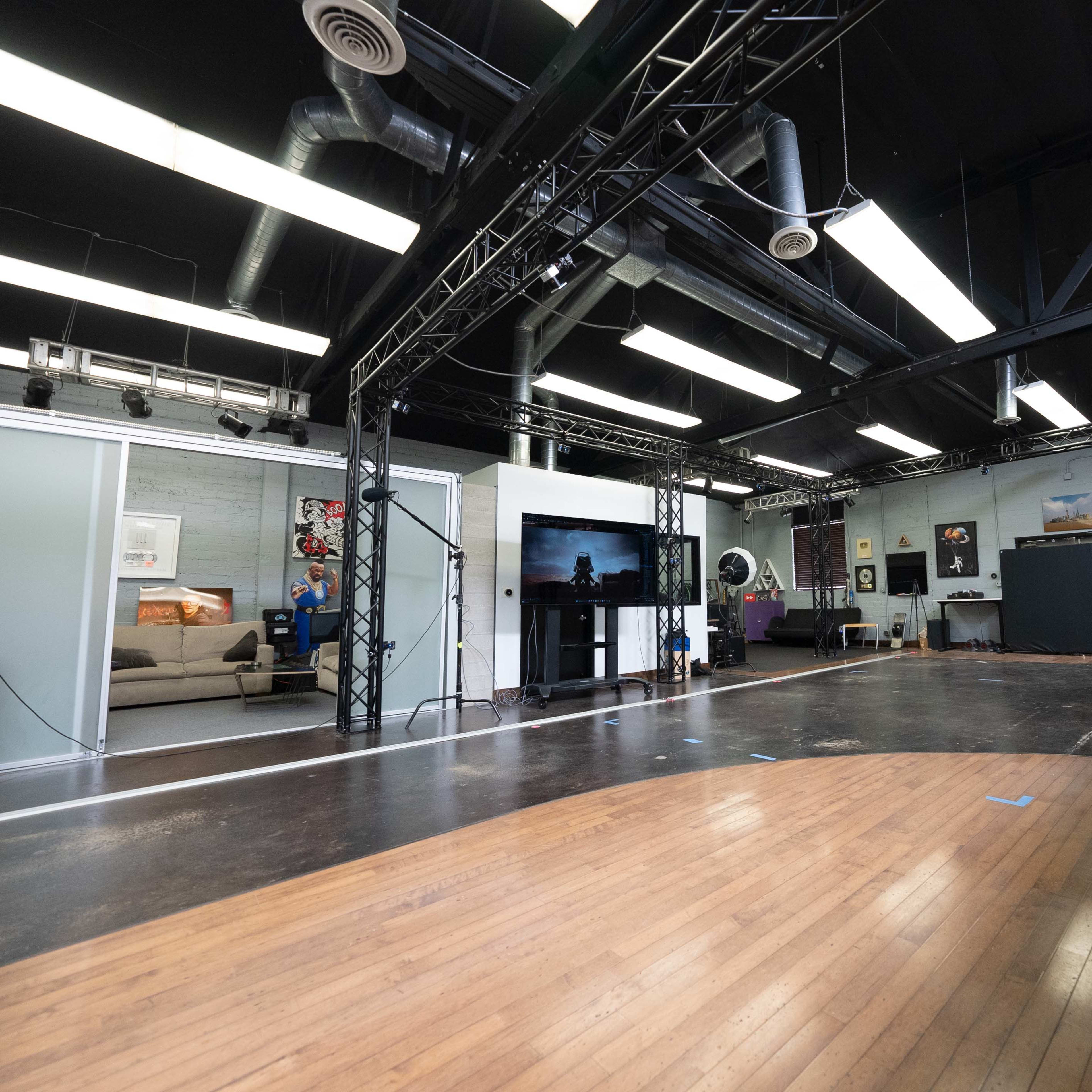 A spacious studio with a wooden floor, black steel trusses, and various decorative elements, including a television and artwork on the walls.