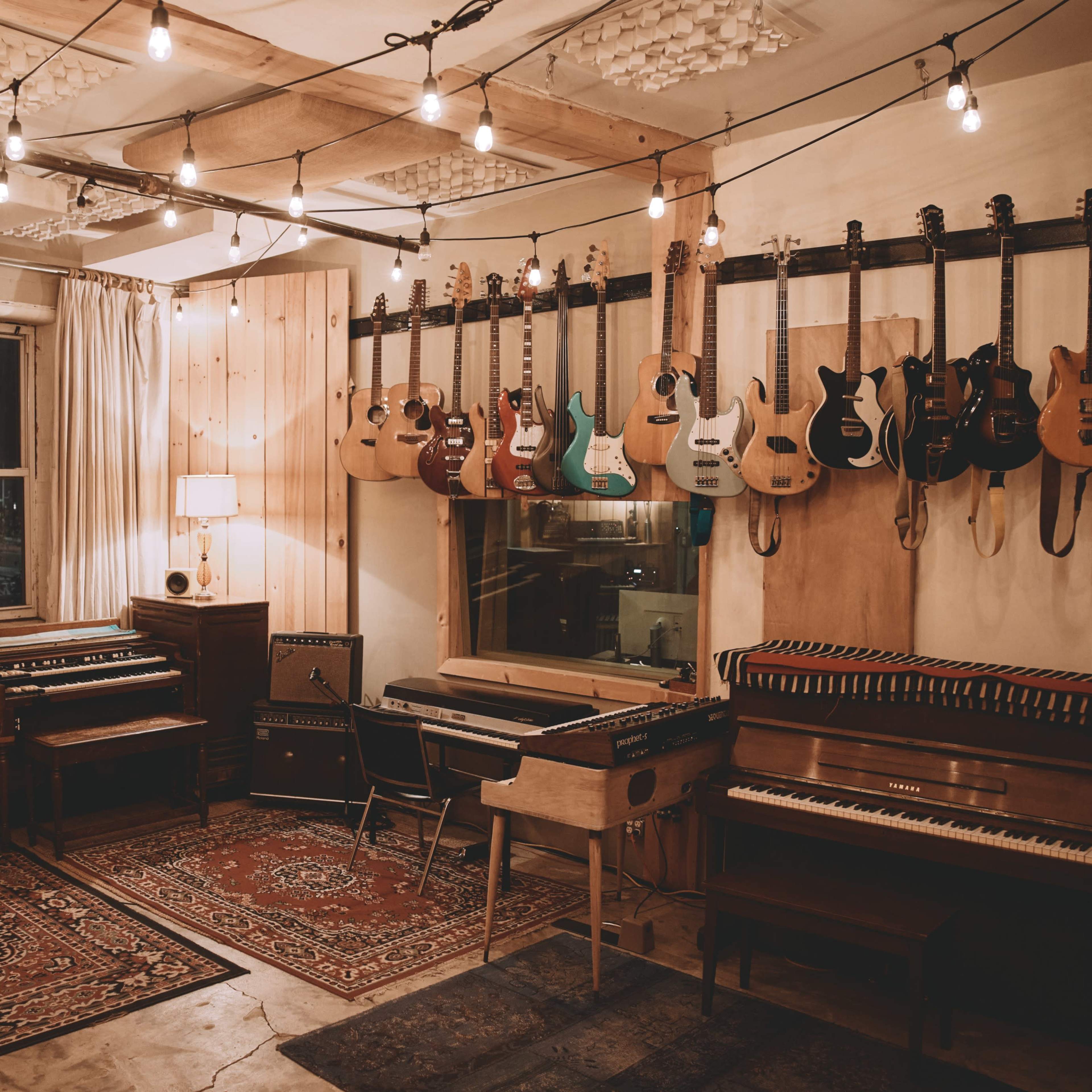 A well-equipped music studio features various guitars hanging on the wall, a vintage piano, and amplifiers arranged around a patterned rug under string lights.
