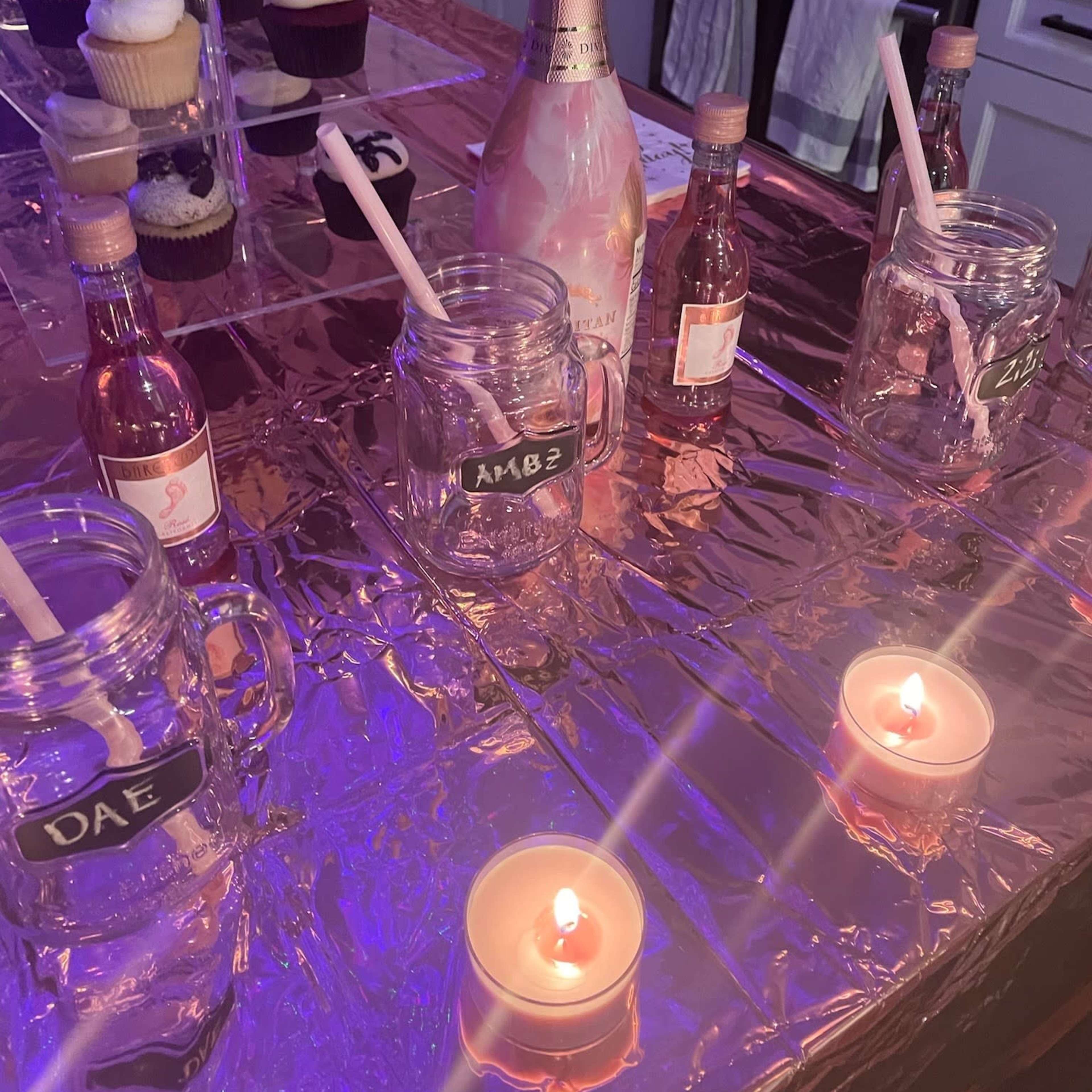 A kitchen countertop decorated with pink champagne, jars filled with colorful liquids, cupcakes, and lit candles.