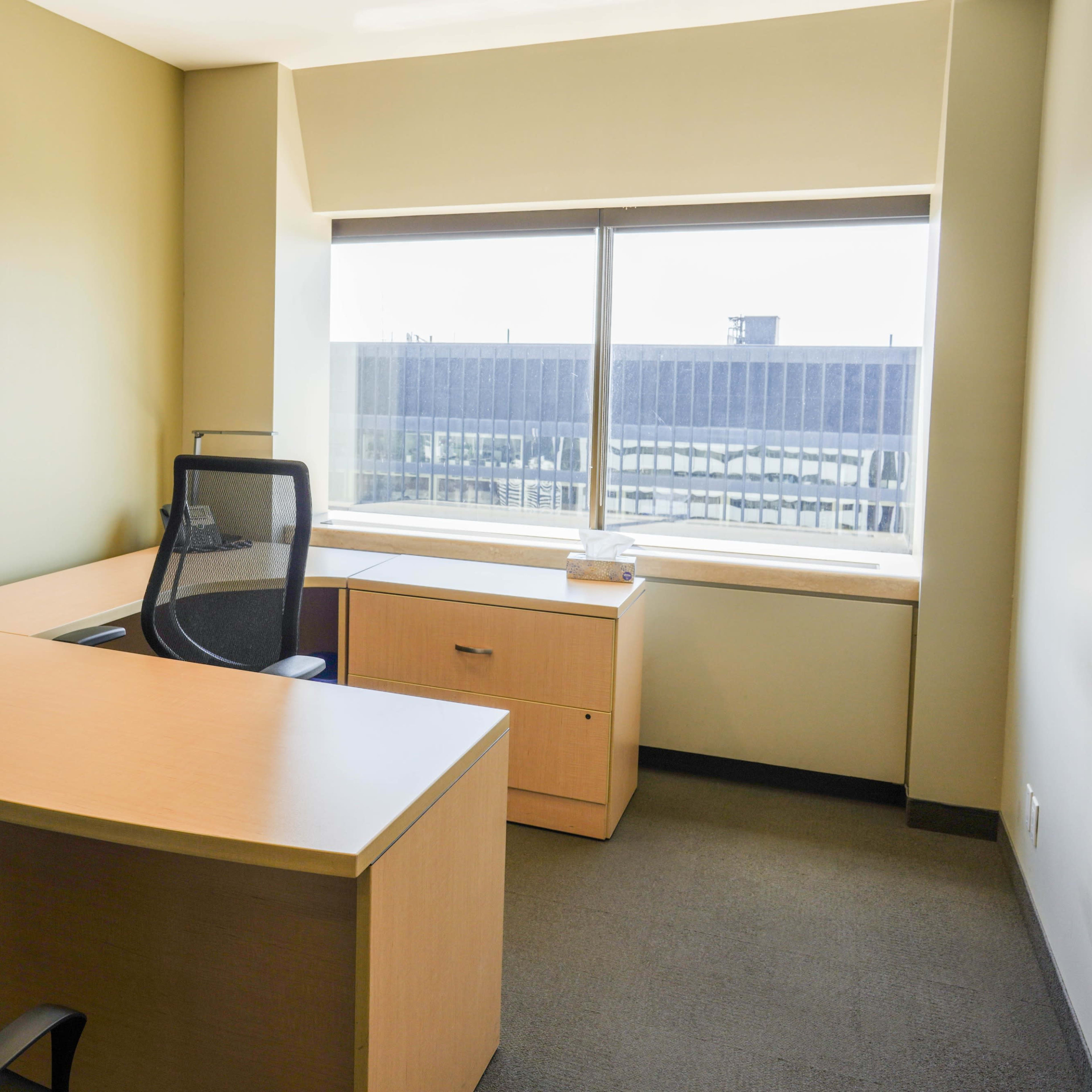 An office space features a wooden desk, a black ergonomic chair, and a large window with a view.