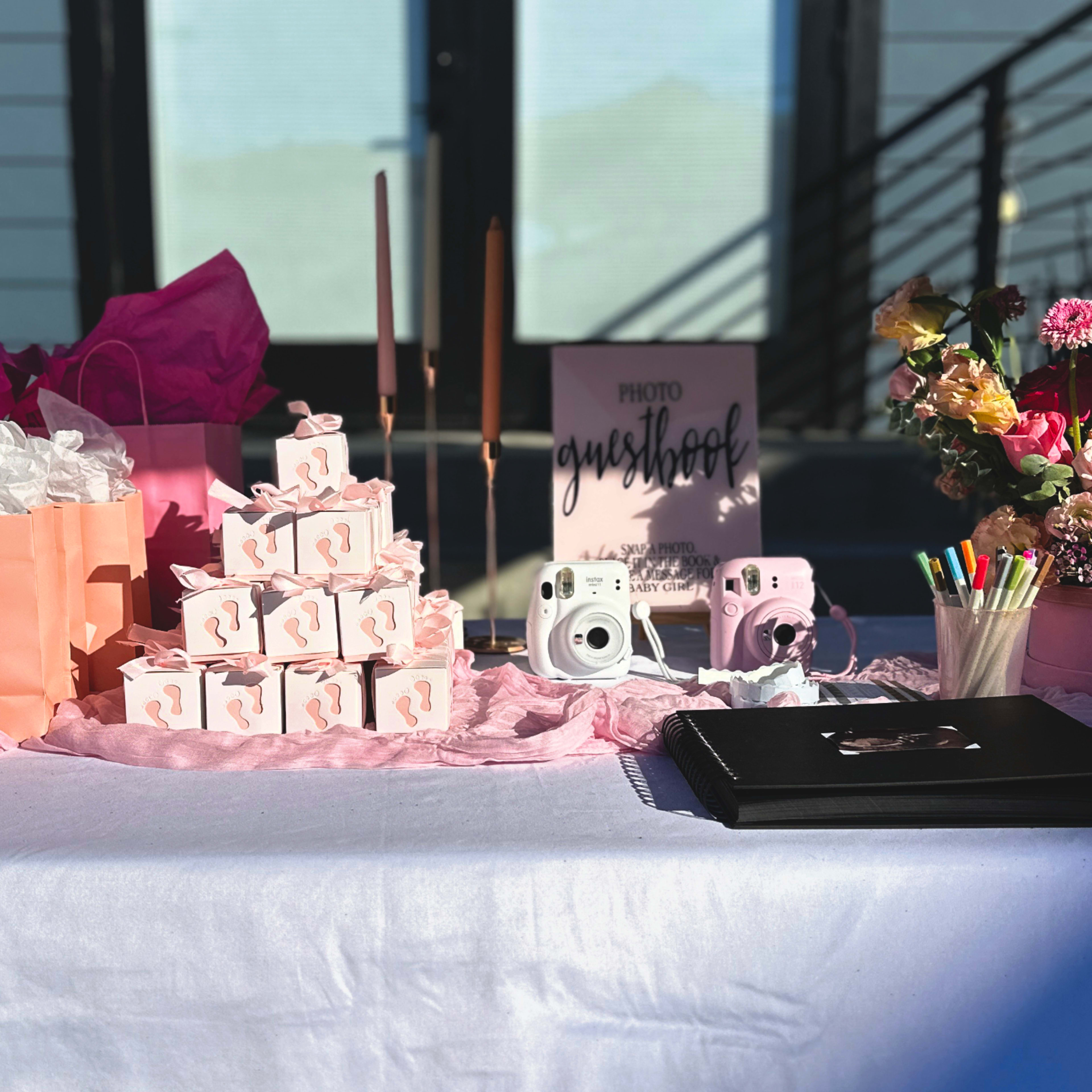 A table is decorated with pink gift bags, stacked boxes, a photo booth sign, floral arrangements, and supplies for a photography session.