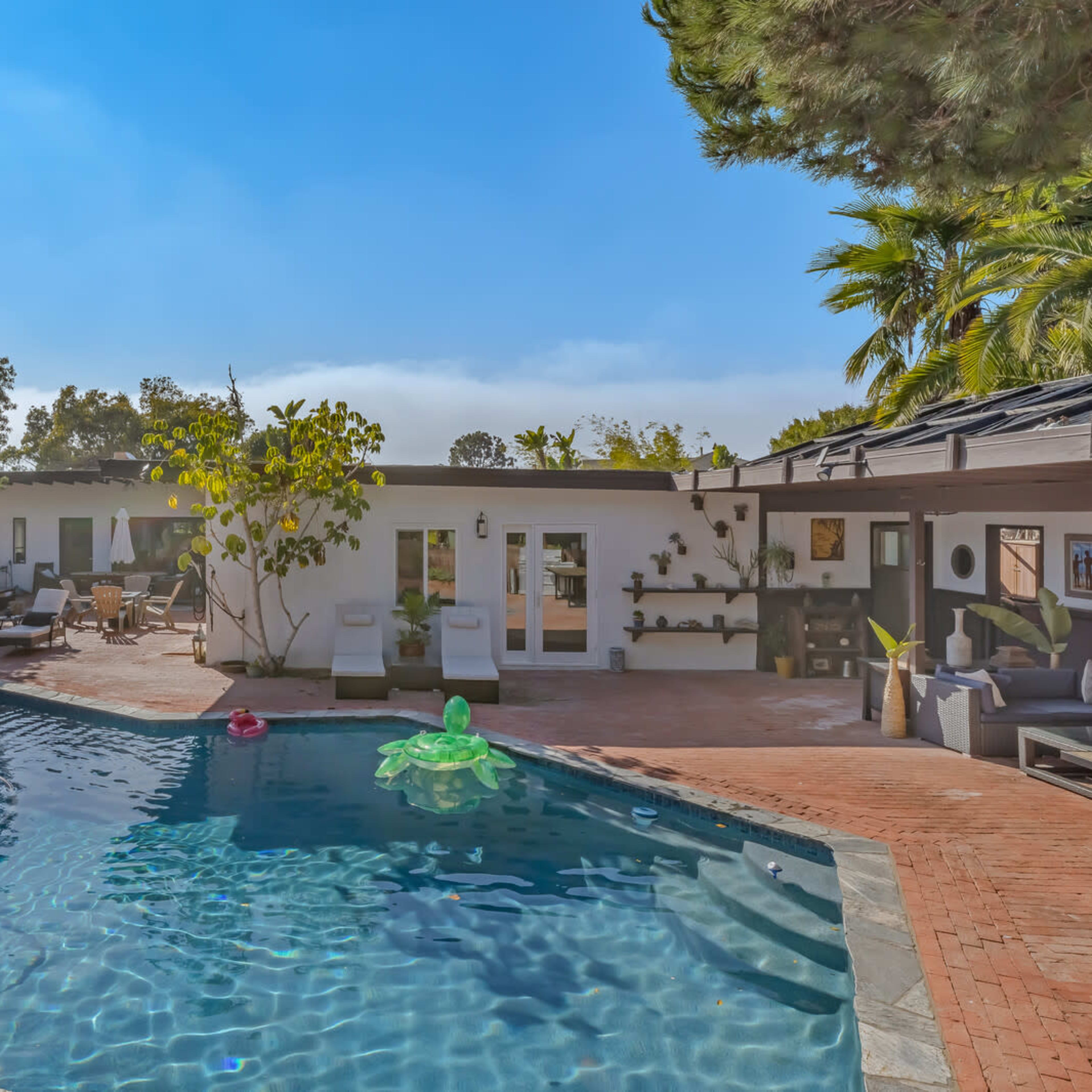 A spacious swimming pool surrounded by lounge chairs and greenery, adjacent to a single-story house with a patio area.