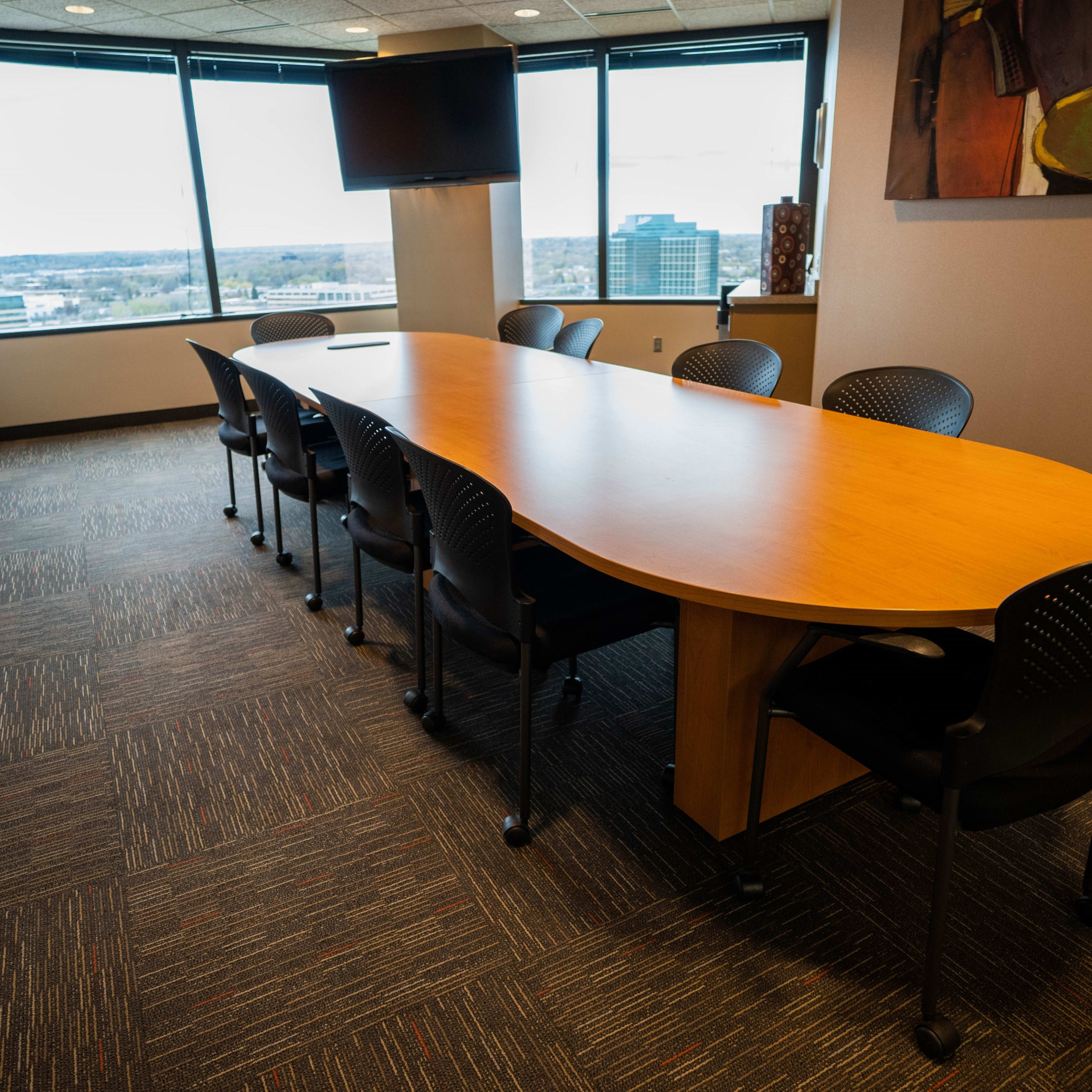 A long, oval conference table with black chairs is positioned in a modern meeting room surrounded by large windows.