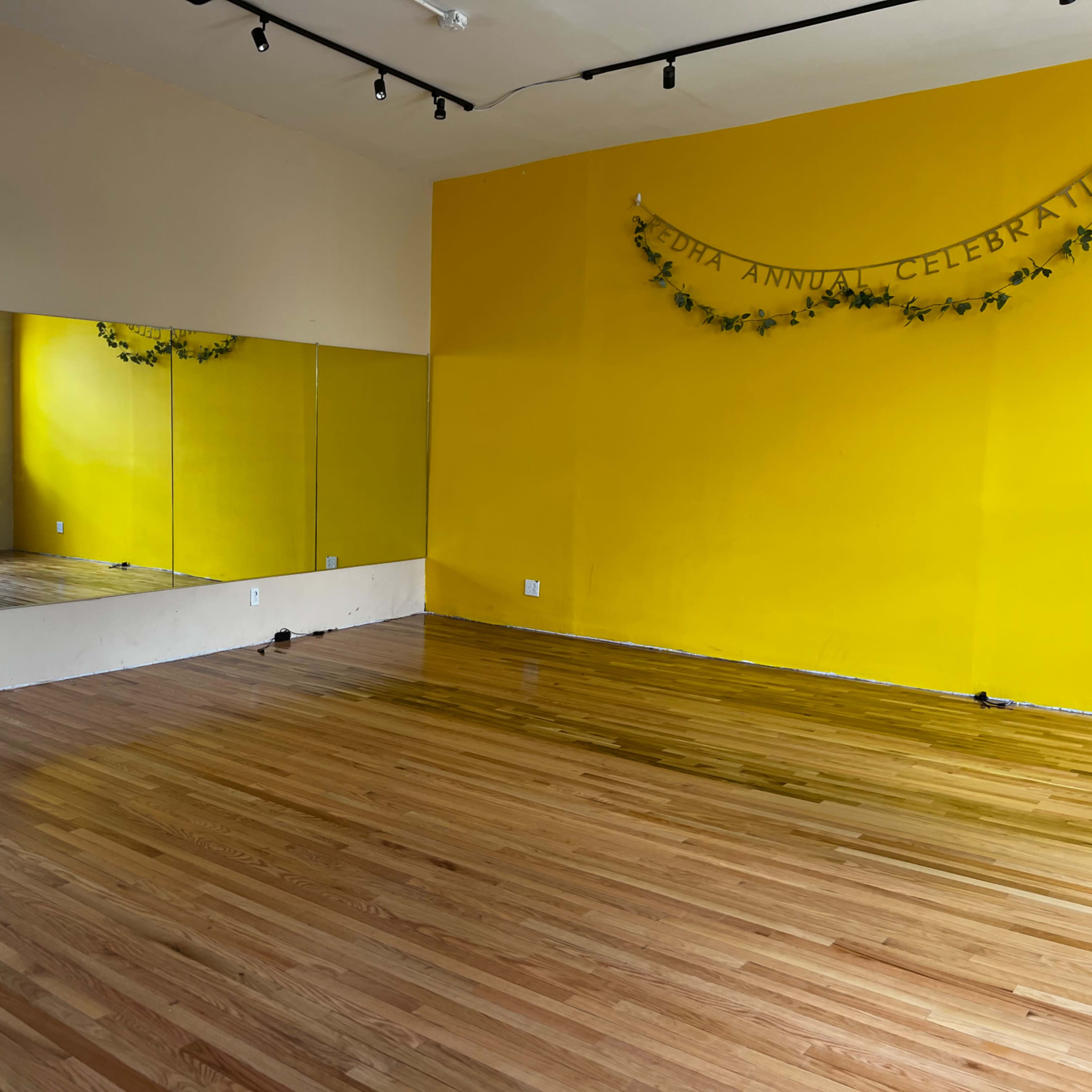 A spacious studio with yellow walls, a wooden floor, and a large mirror along one side.