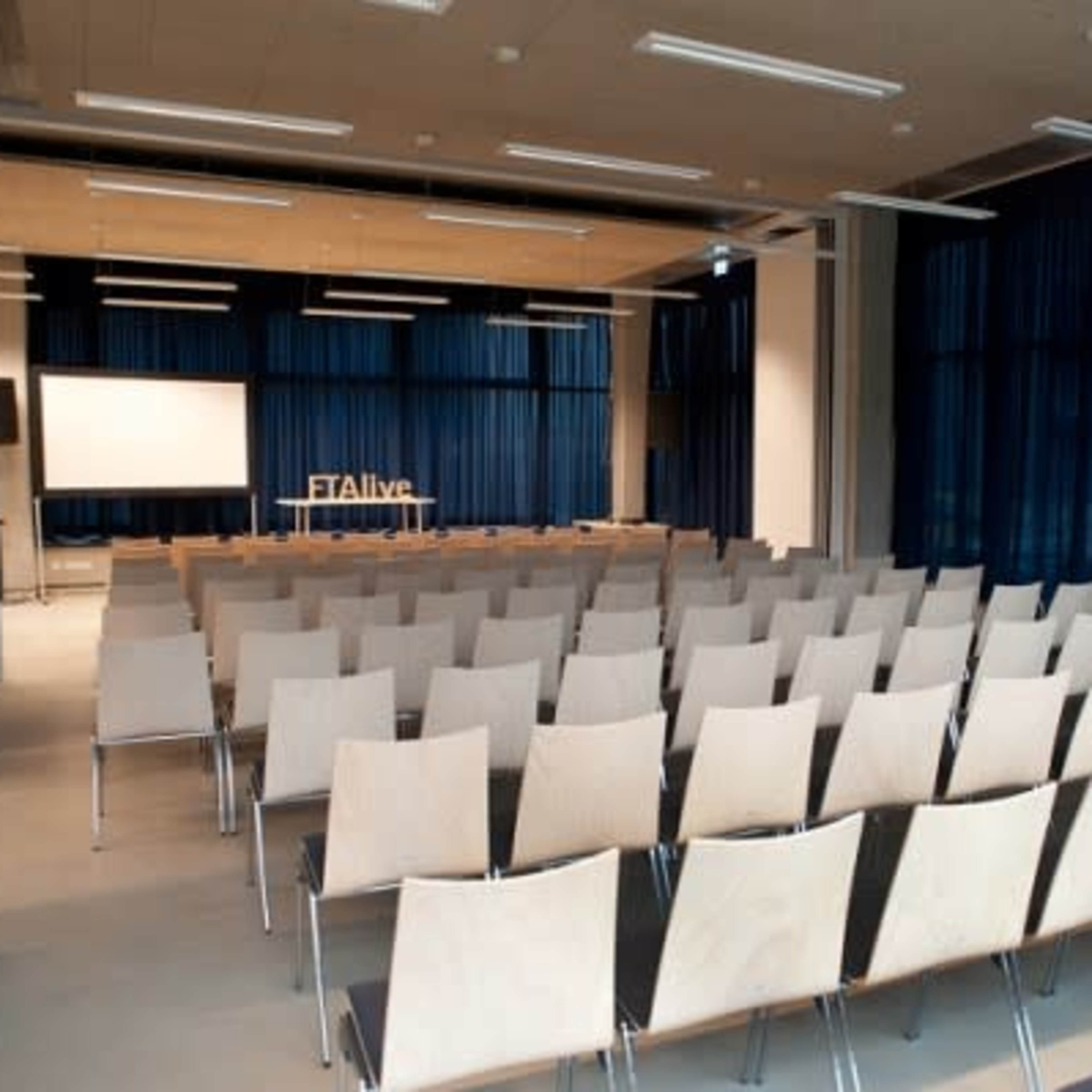A spacious conference room with rows of empty wooden chairs facing a large screen and a backdrop with the text "#Alive."