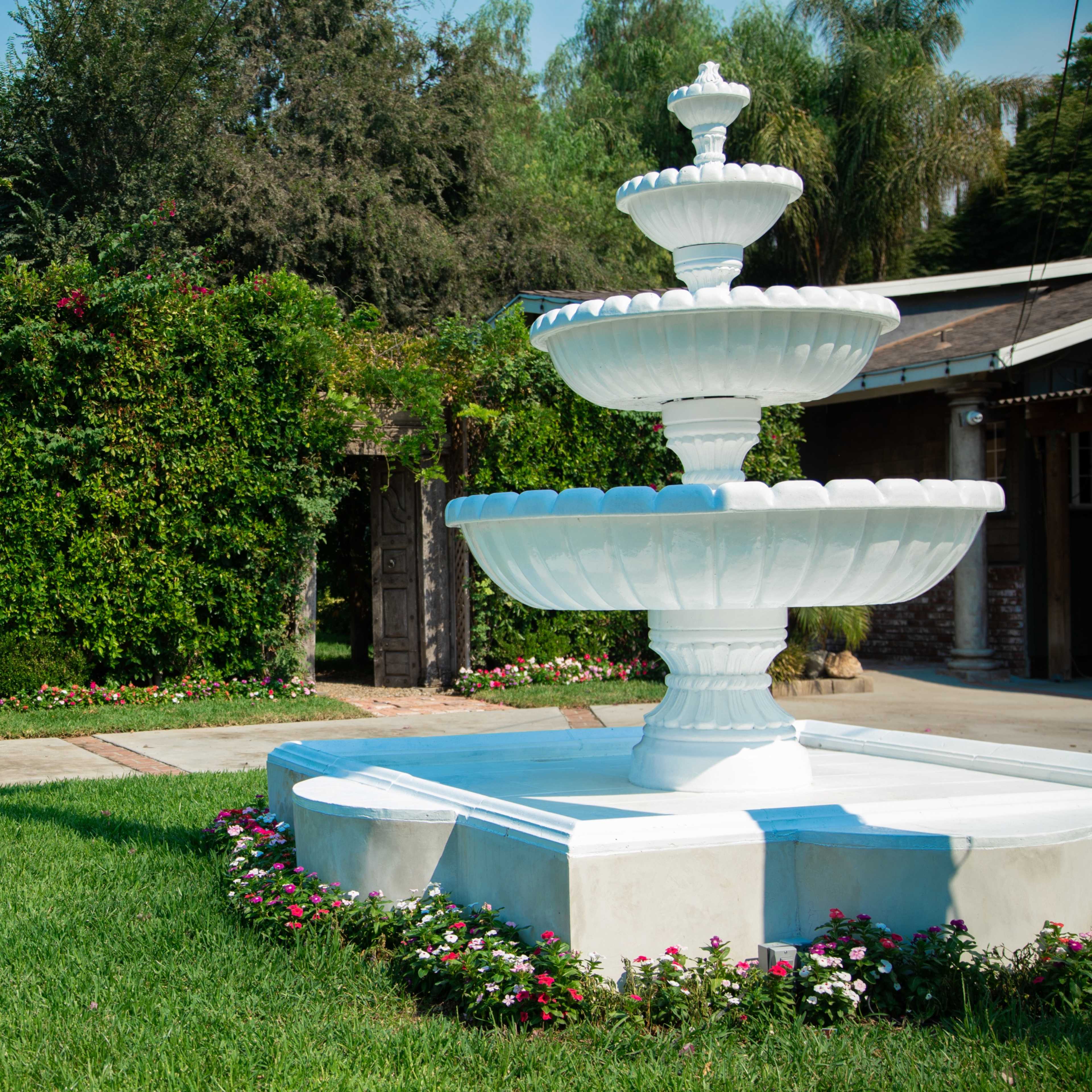 A white three-tiered fountain is located in a grassy area surrounded by vibrant greenery and flower beds.