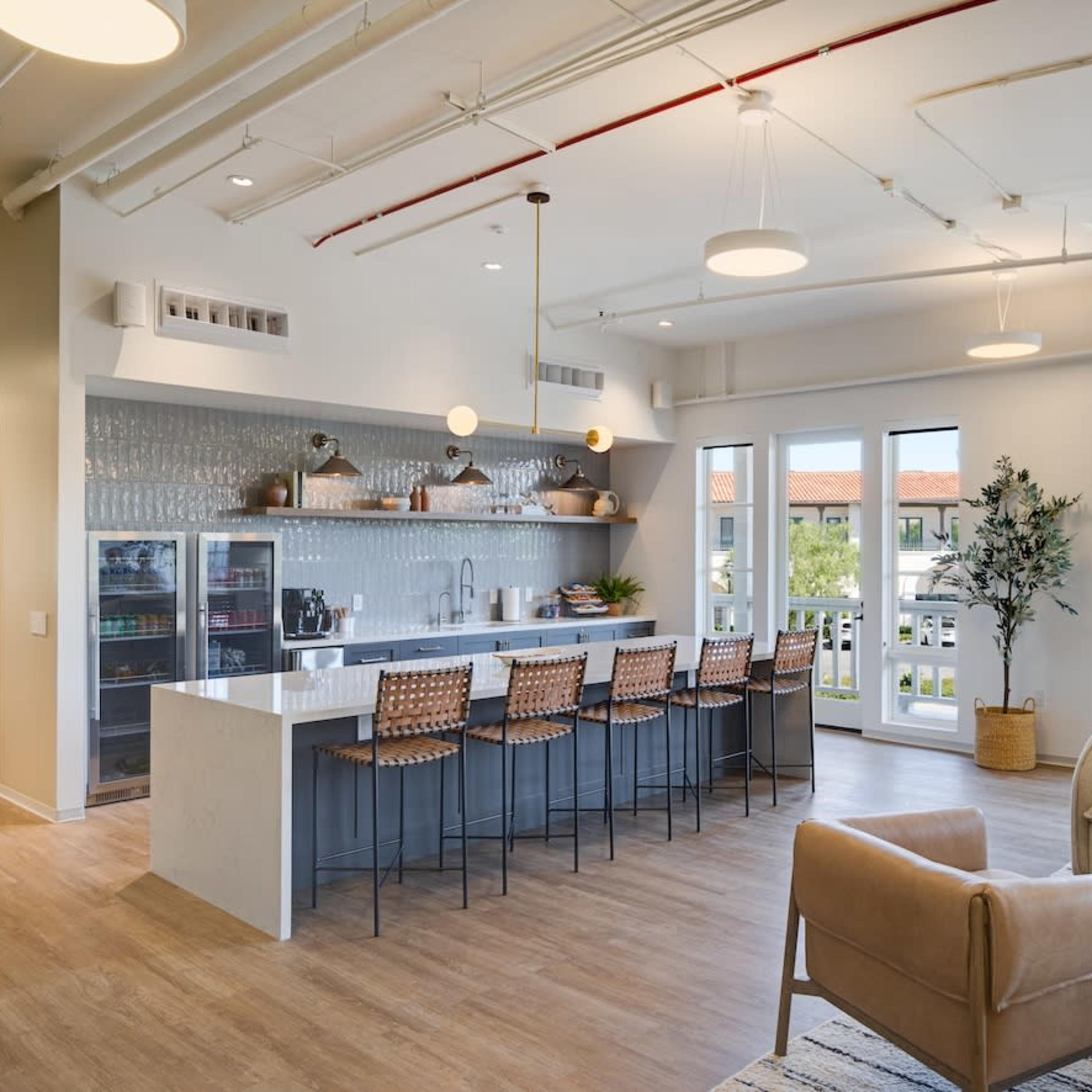 A modern kitchen area with a long countertop, high chairs, and large windows allowing natural light to illuminate the space.