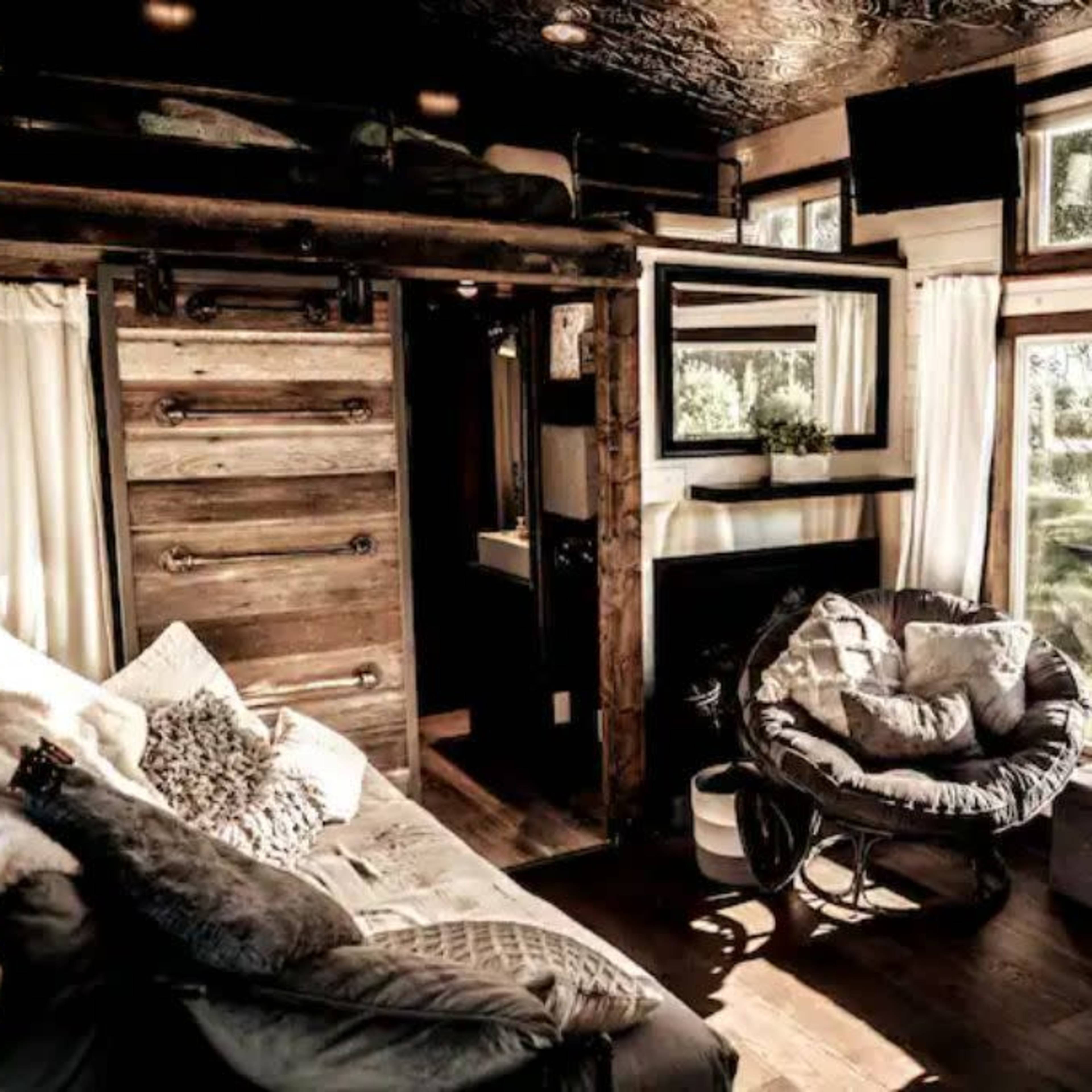 A cozy interior of a tiny house featuring a sitting area with a large window overlooking greenery, decorated with plush seating and wooden accents.