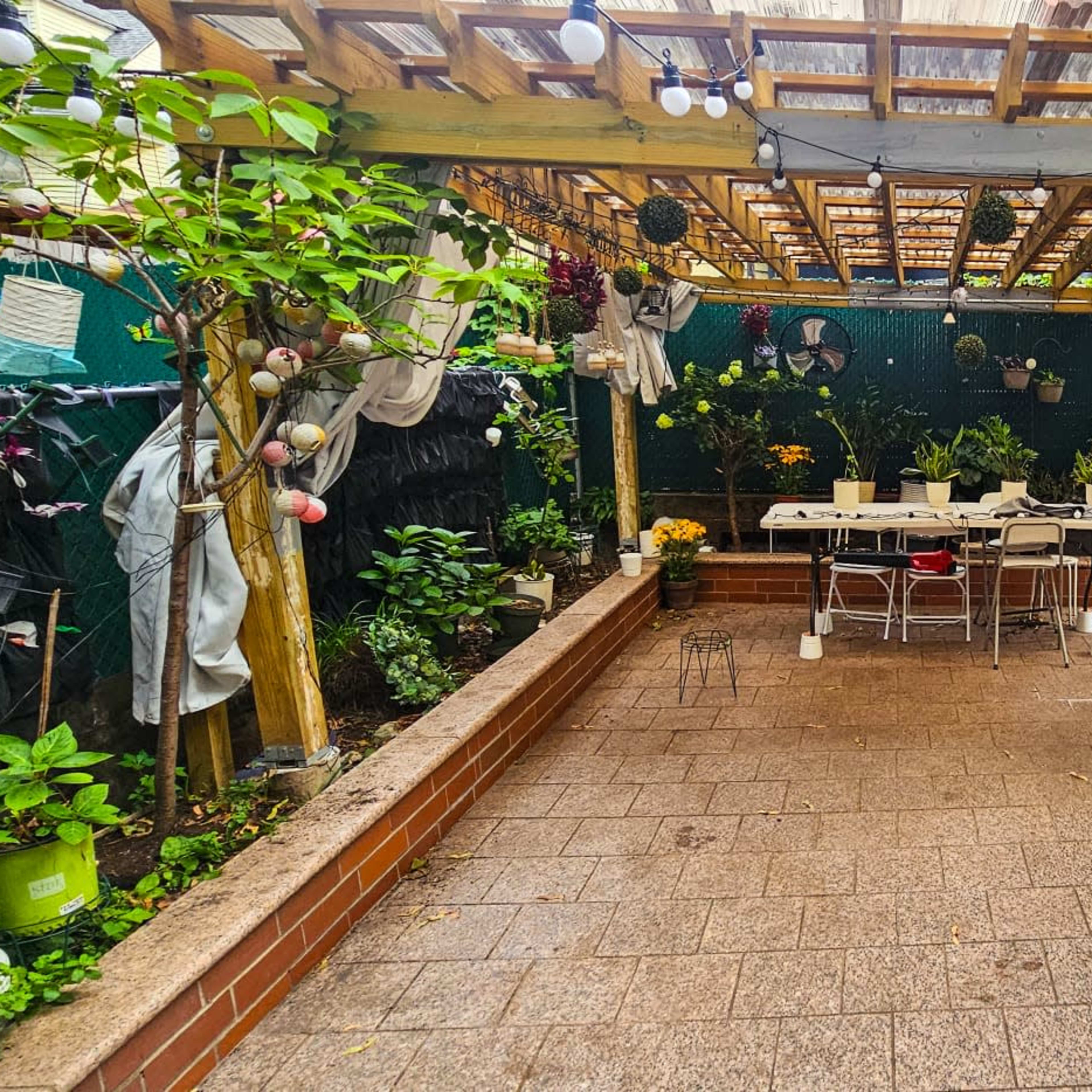A landscaped patio area with a wooden pergola, a dining table, chairs, and various potted plants arranged around the space.