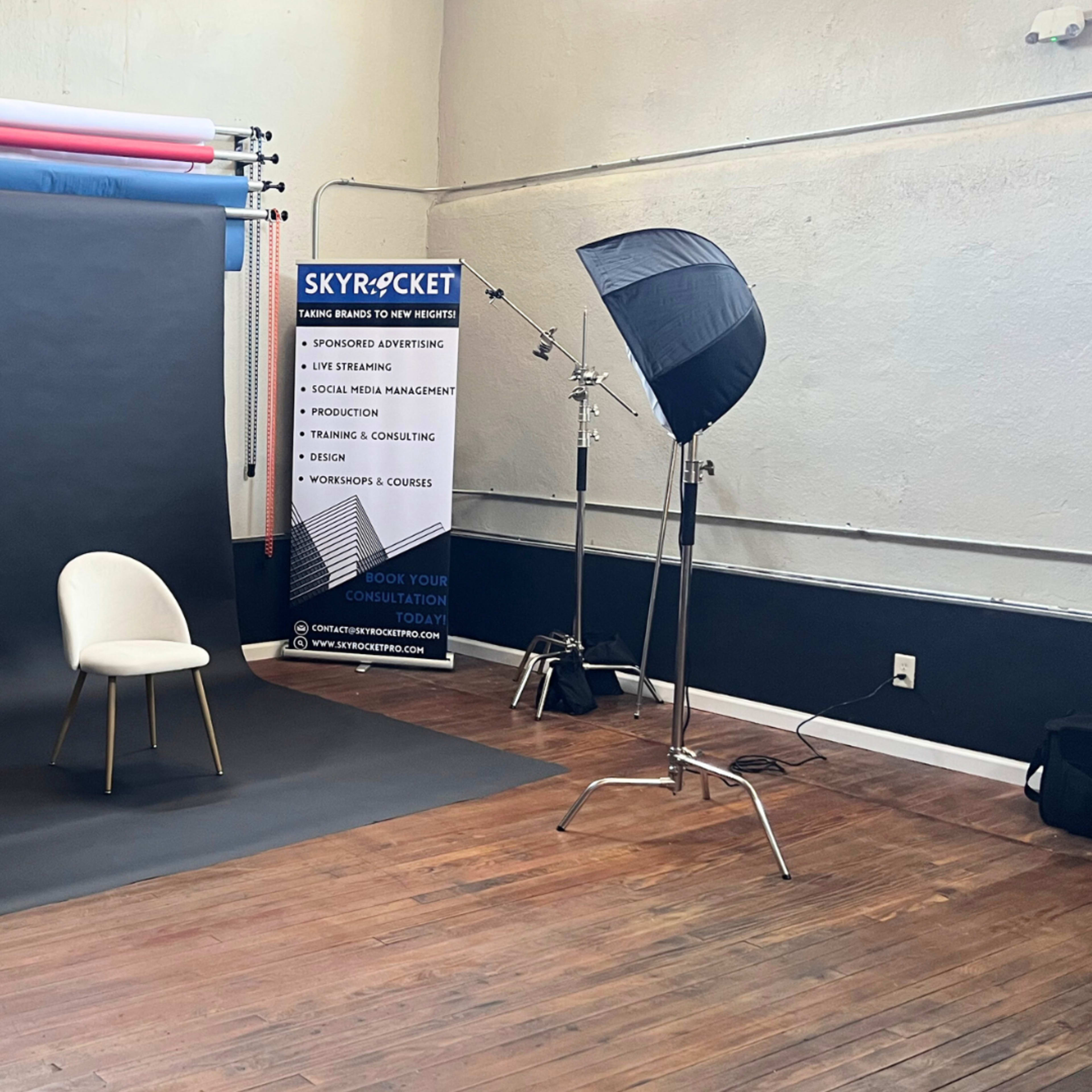 A photography studio setup with a black backdrop, a single chair, and a light source on a stand, near a promotional banner.
