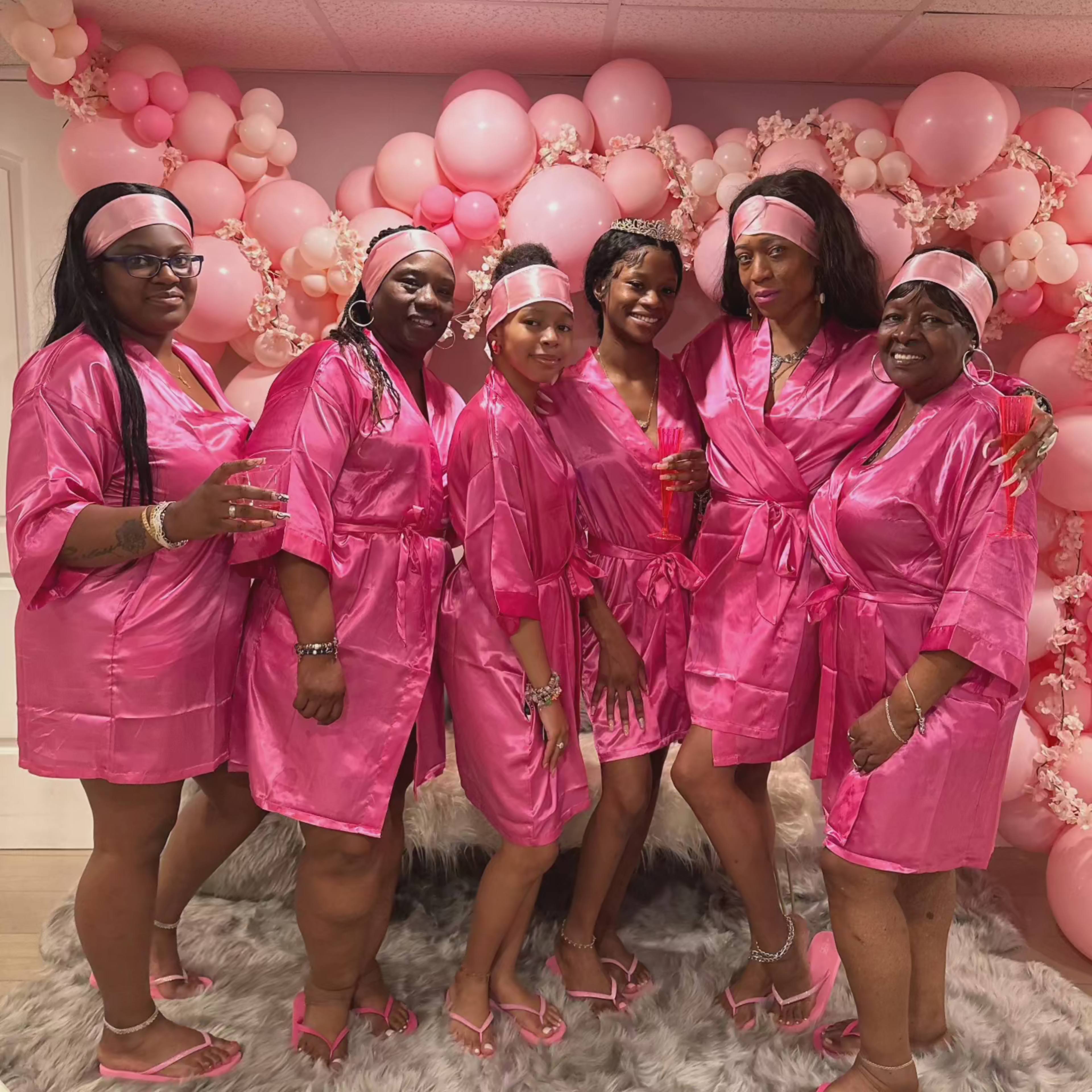 A group of six women in matching pink satin robes stands in front of a decorative backdrop of pink balloons.