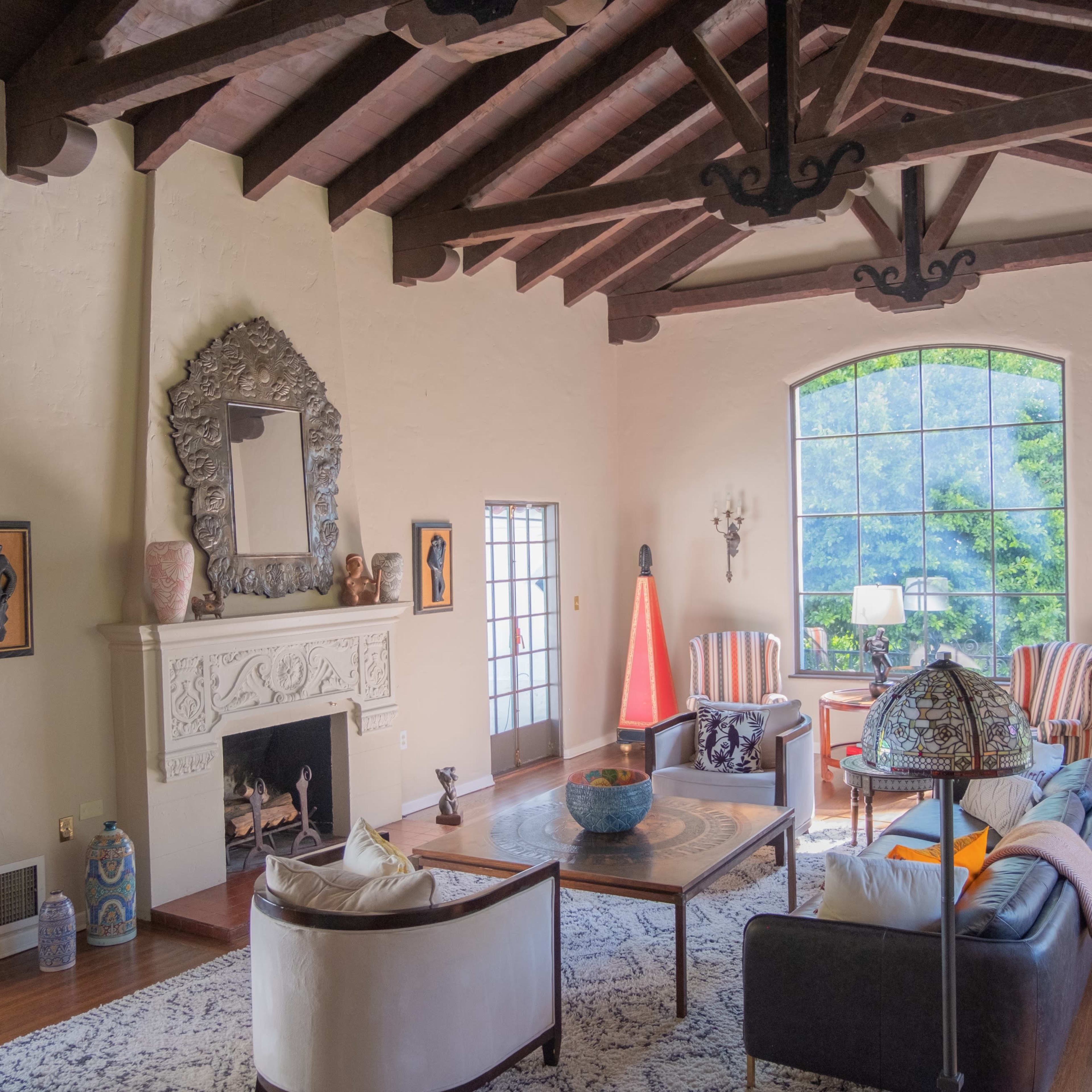 A spacious living room featuring a high ceiling with wooden beams, a fireplace, large windows, and various decorative elements including artwork and furniture.