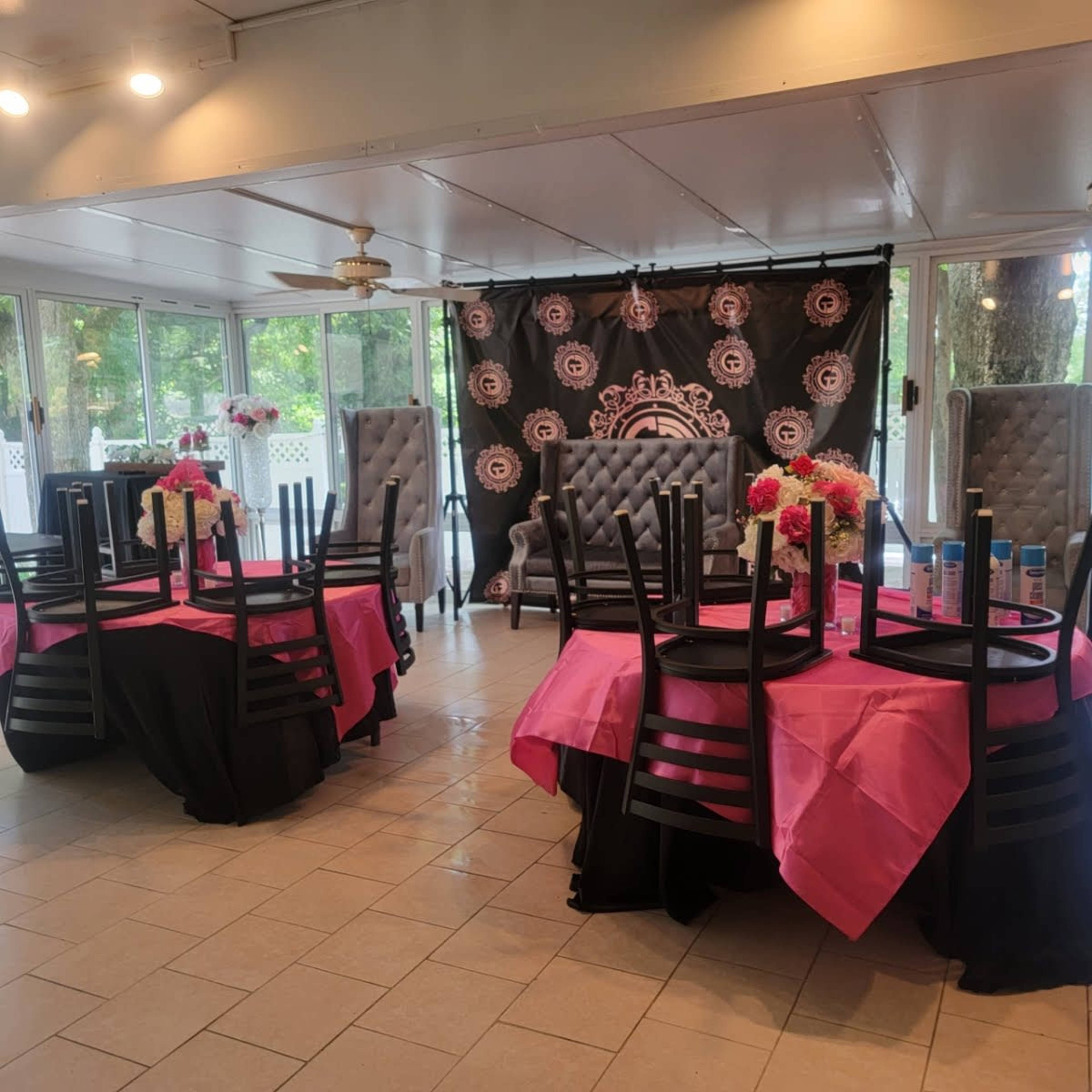 A banquet room with decorated tables covered in pink and black tablecloths, surrounded by chairs stacked upside down, and a backdrop featuring a patterned design.