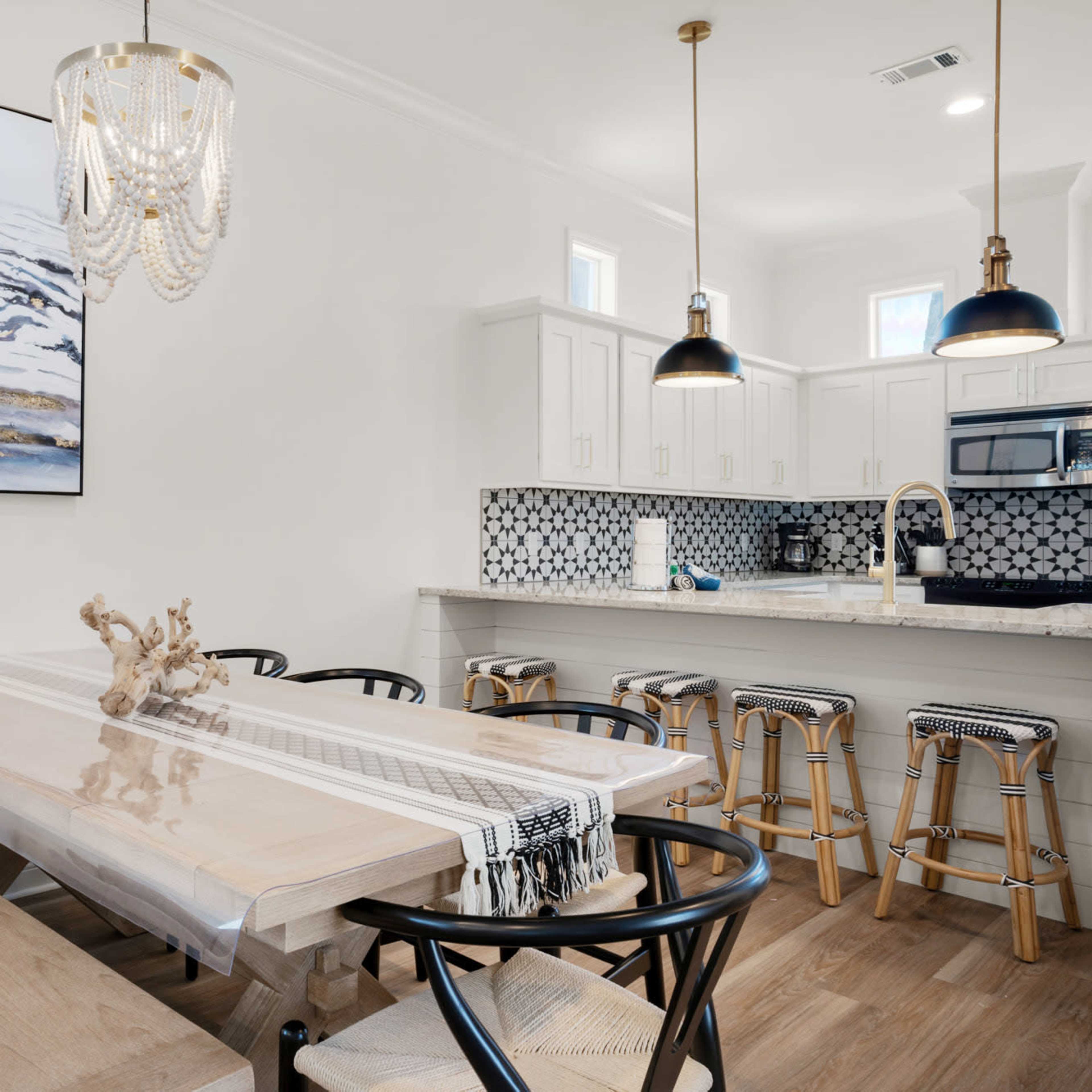 A modern dining area features a long wooden table with a decorative centerpiece, surrounded by sleek black chairs, adjacent to a kitchen with white cabinetry and a patterned tile backsplash.