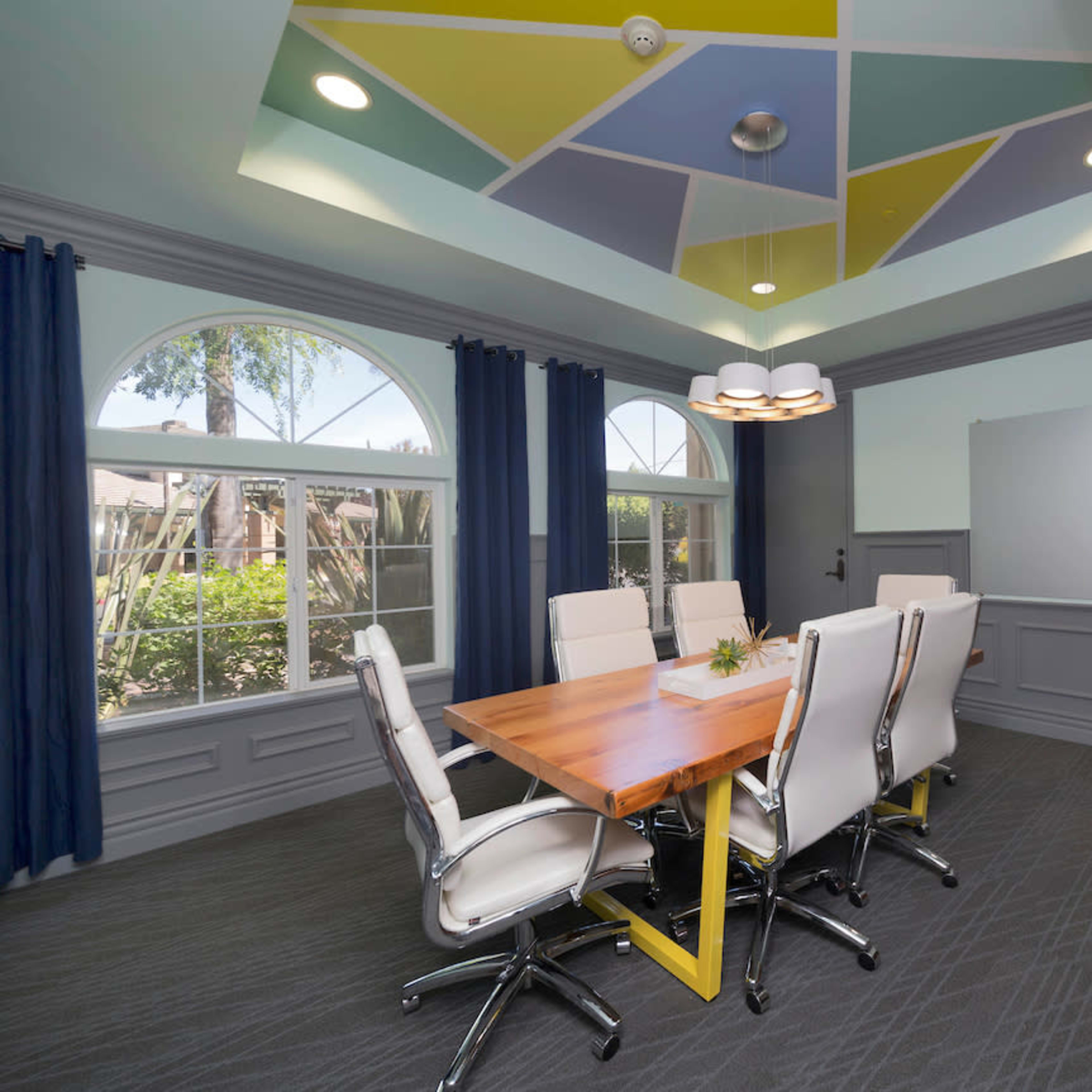 A conference room features a wooden table surrounded by white chairs, with large windows offering a view of greenery and a ceiling adorned with colorful geometric patterns.