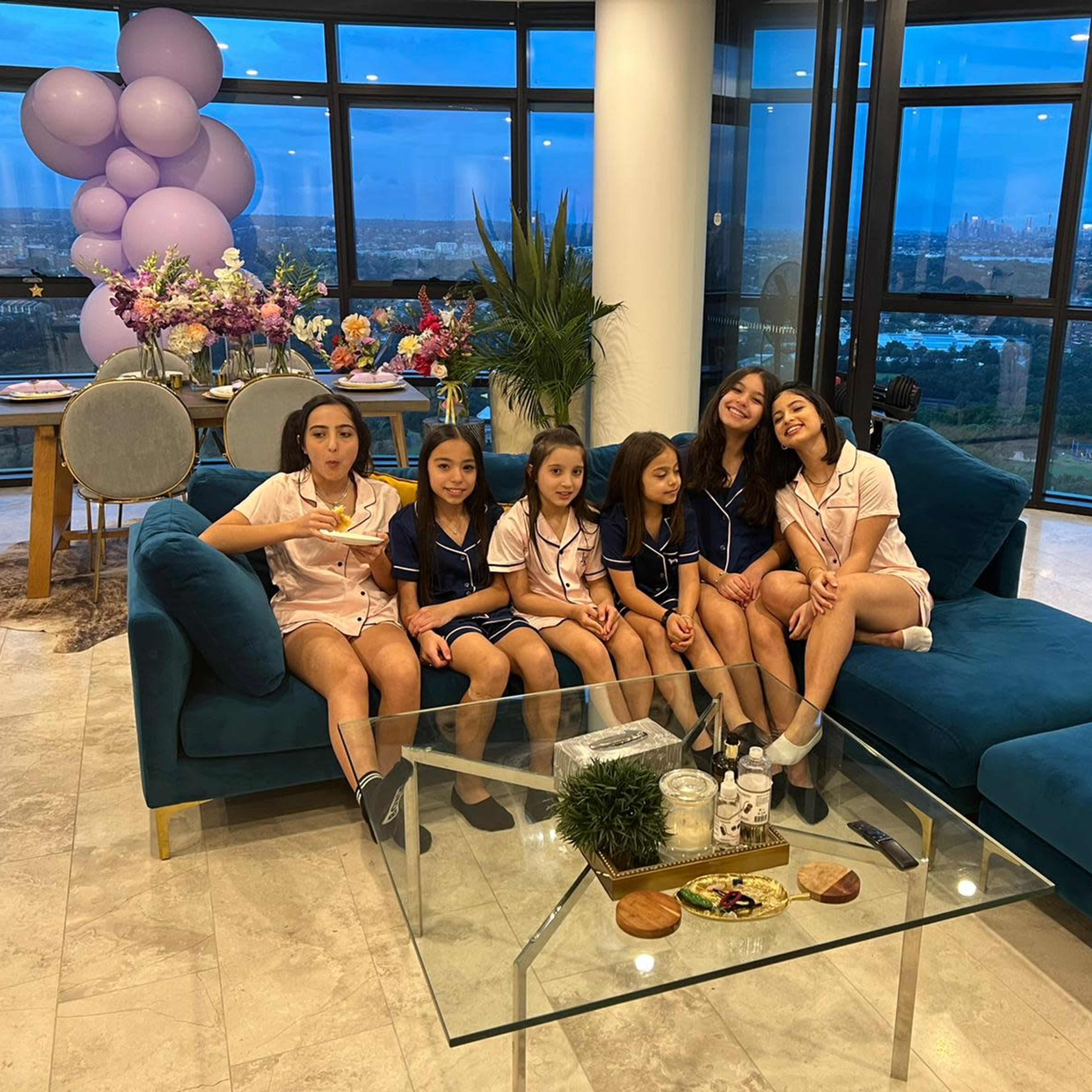 A group of six children in matching pajamas poses smiling on a sofa in a bright, modern living room with large windows and decorative elements.