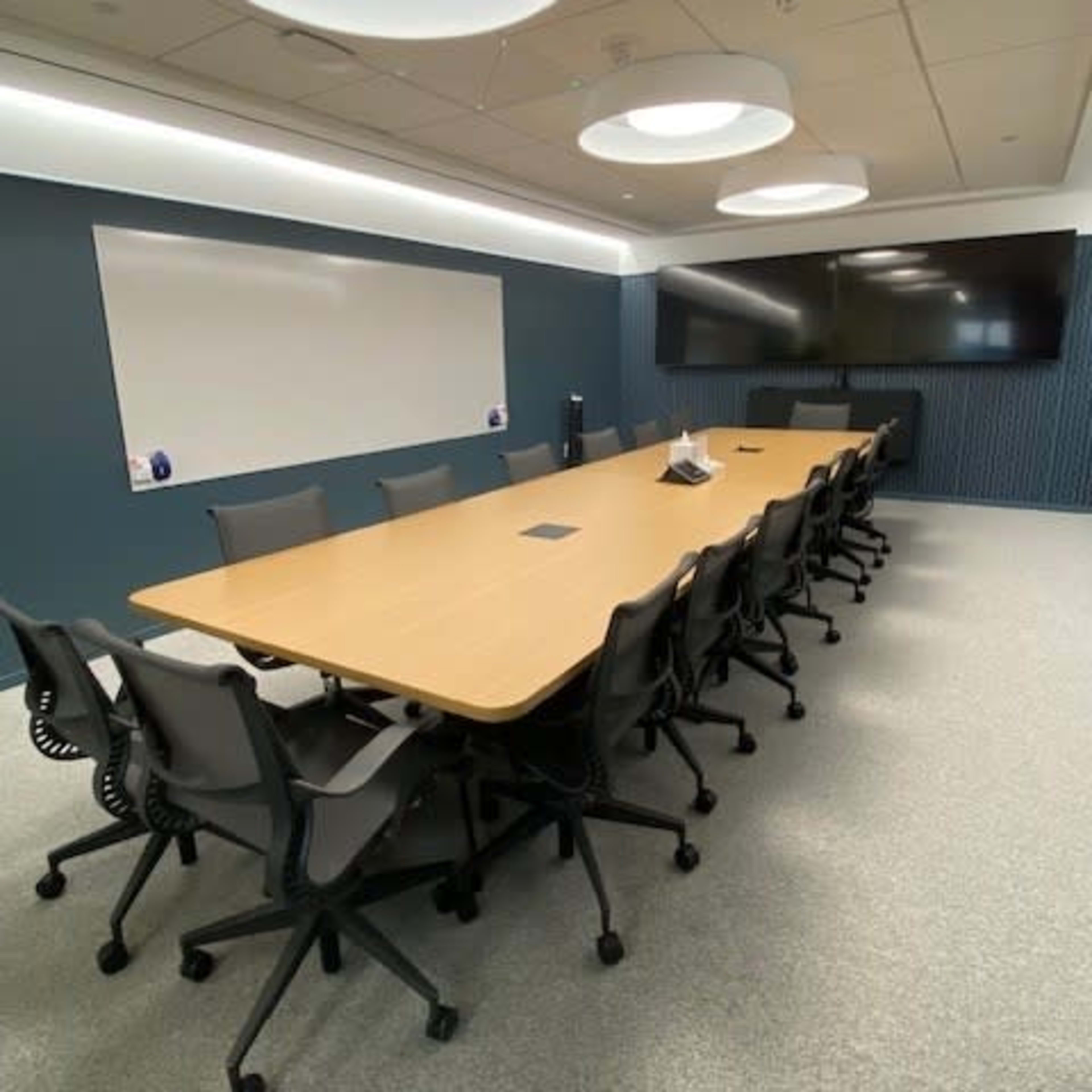 A modern conference room with a long wooden table surrounded by ergonomic office chairs, a large screen on the wall, and a whiteboard.