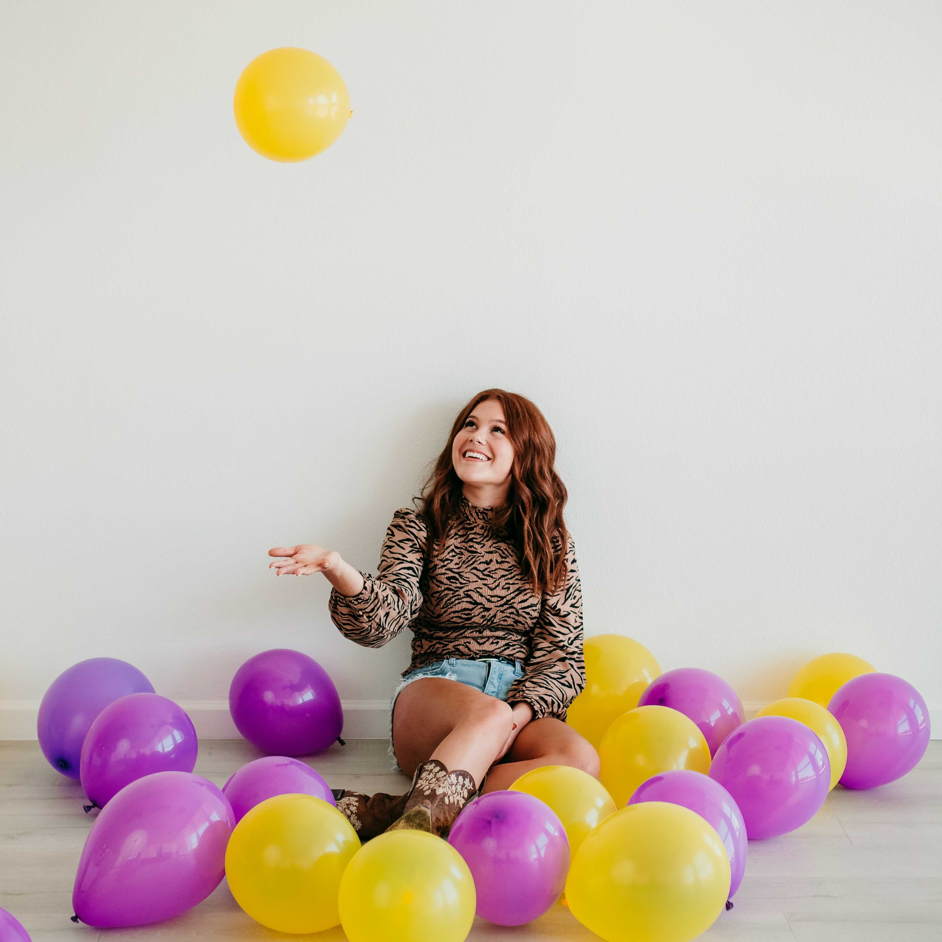 A woman sits on the floor surrounded by purple and yellow balloons, playfully tossing a yellow balloon into the air.