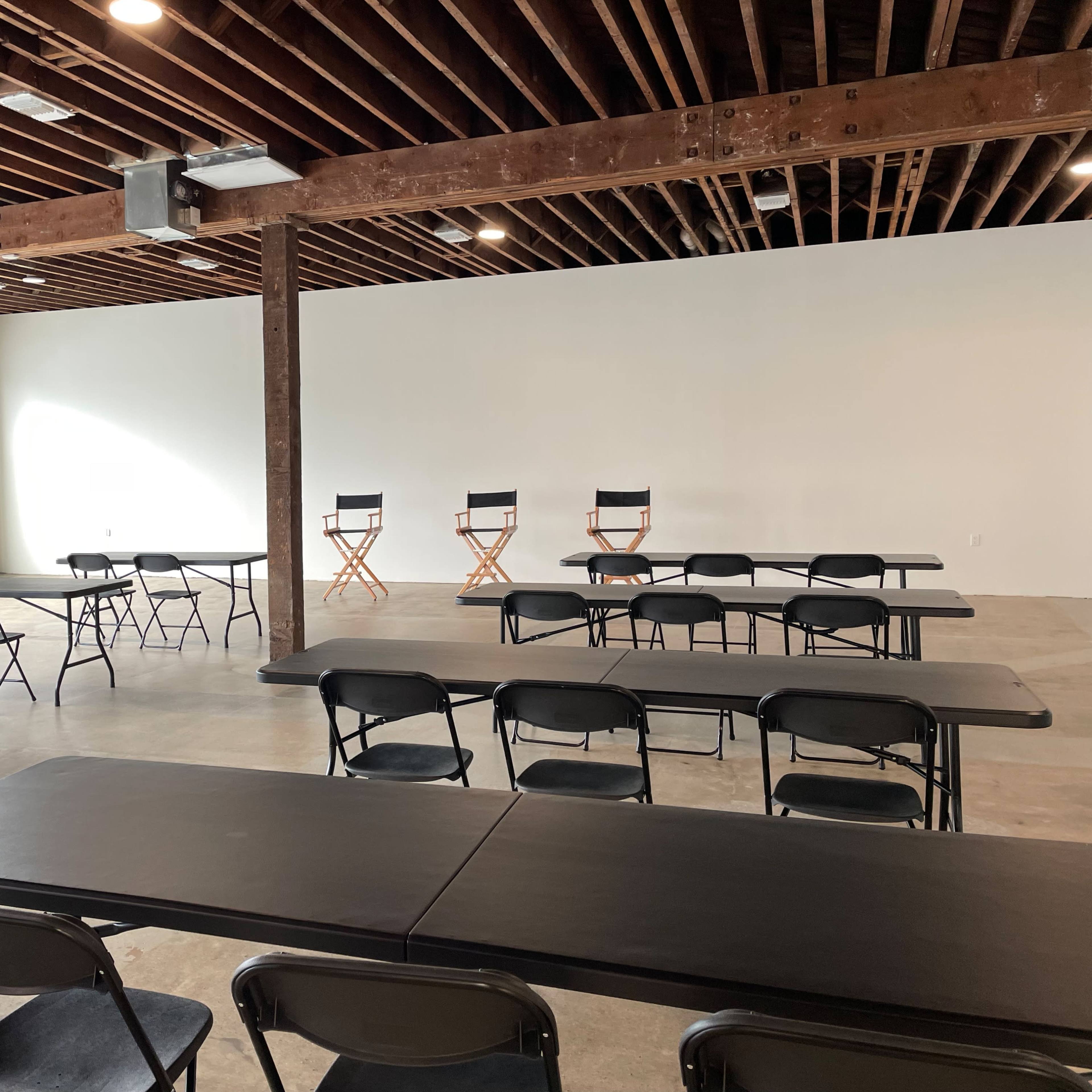 A spacious room with black folding chairs and tables arranged in a grid, featuring three director's chairs positioned against a plain white wall.
