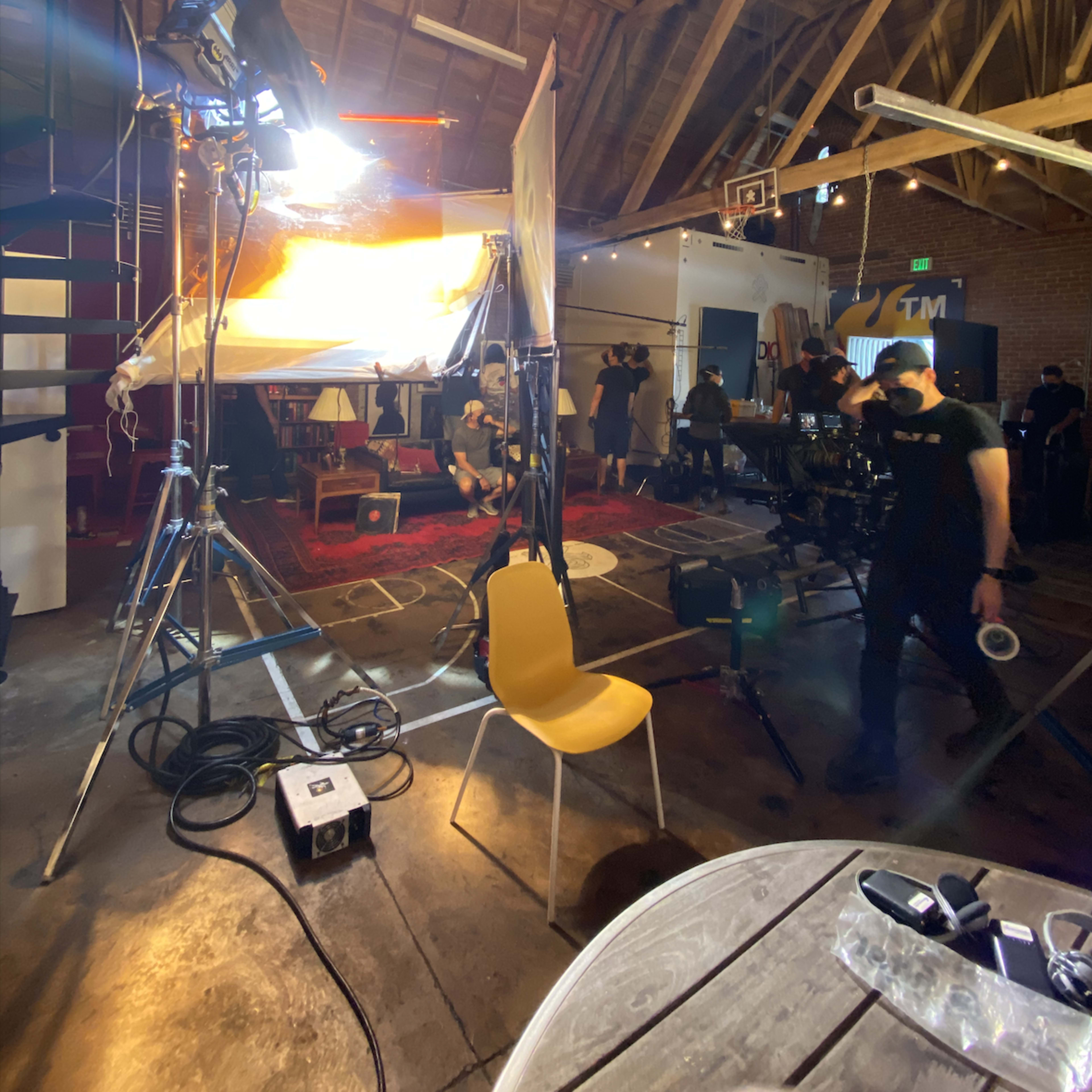 A film crew sets up equipment in a spacious loft with exposed wooden beams and a carpeted area for a video shoot.