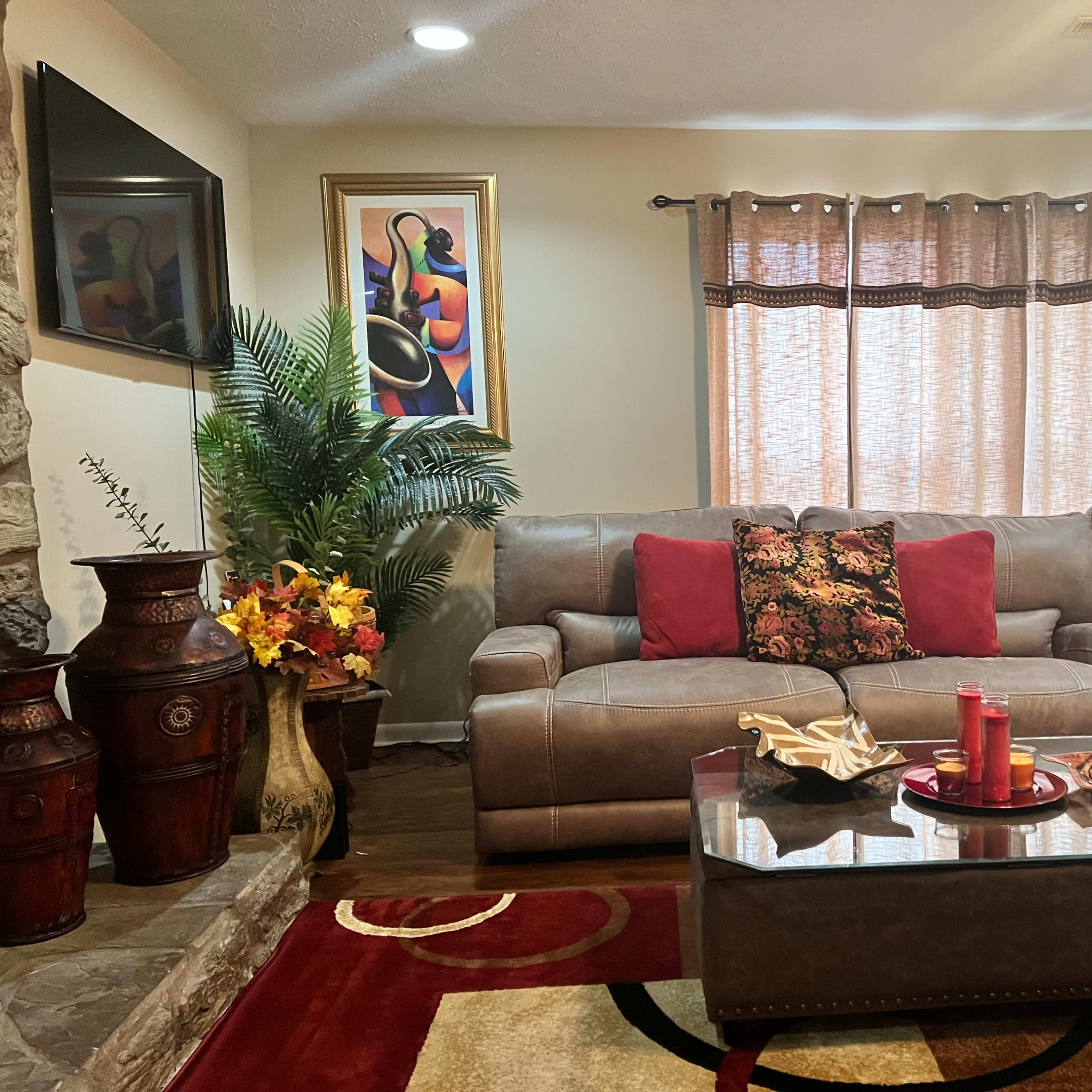 The living room features a beige sofa with red pillows, a glass coffee table, and decorative artwork on the walls, complemented by a potted plant and various decorative items.