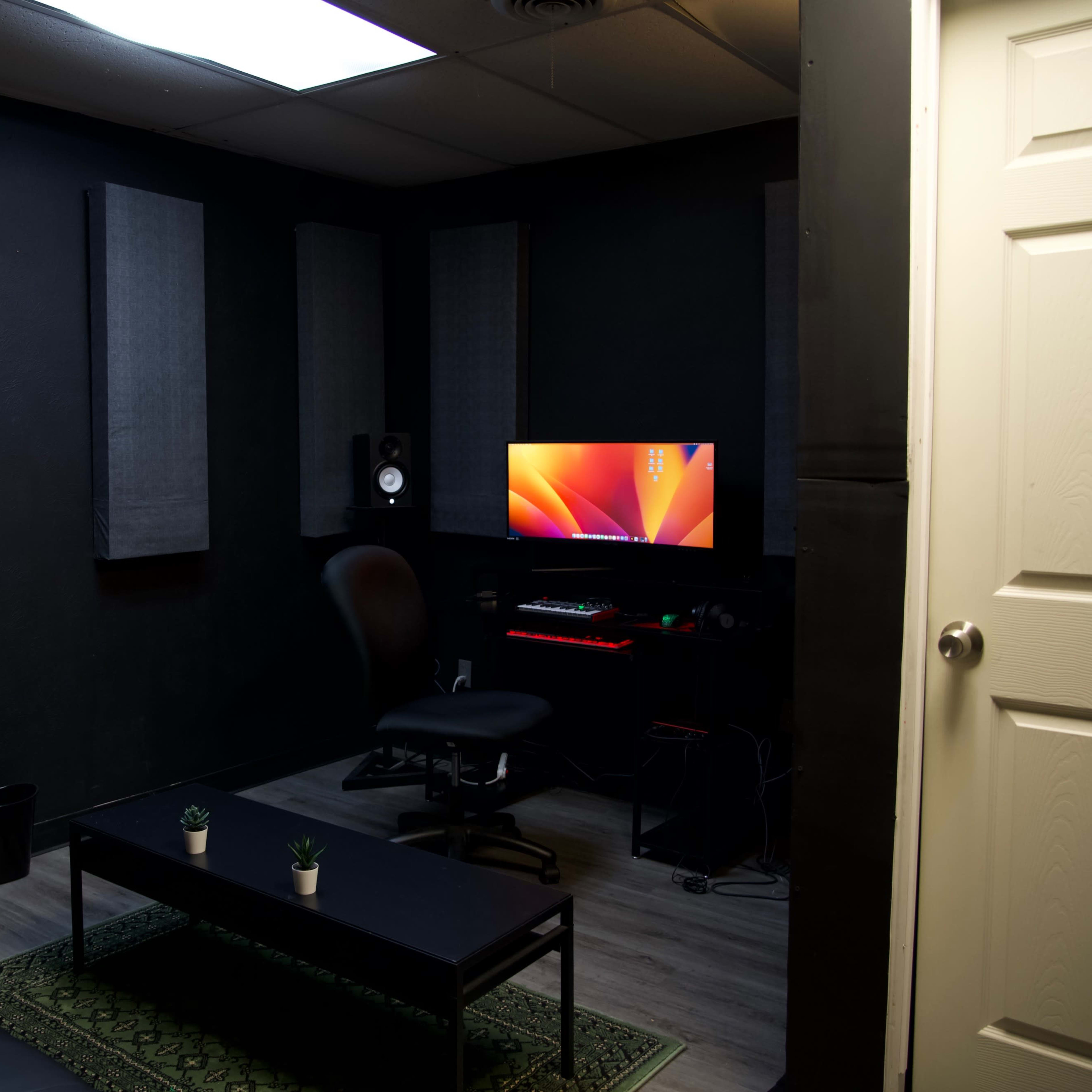 A dimly lit recording studio featuring soundproofing panels on the walls, a modern computer setup on a desk, and a black couch with a coffee table in the center.