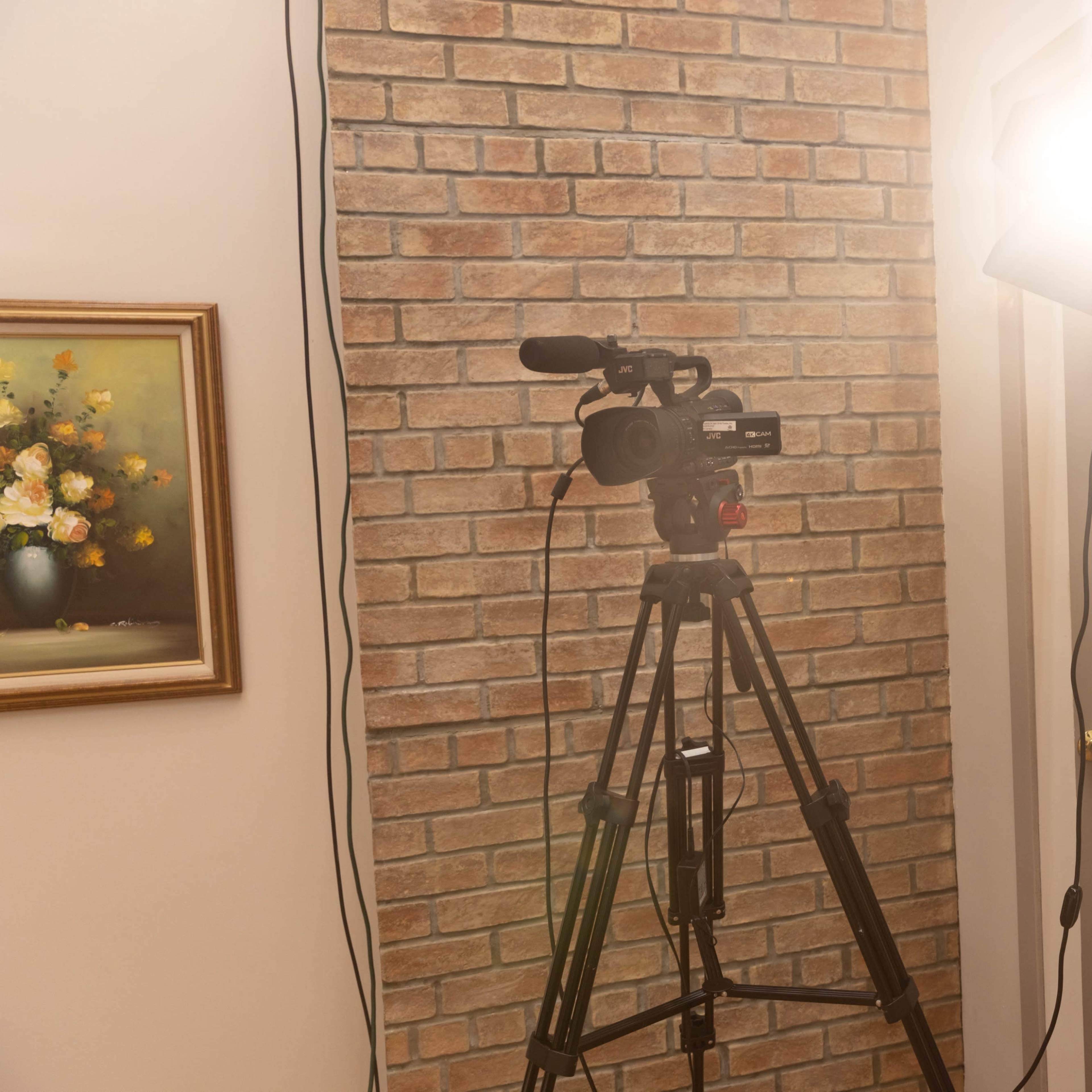 A video camera on a tripod is positioned next to a bright light source in a room with a brick wall and a floral painting.