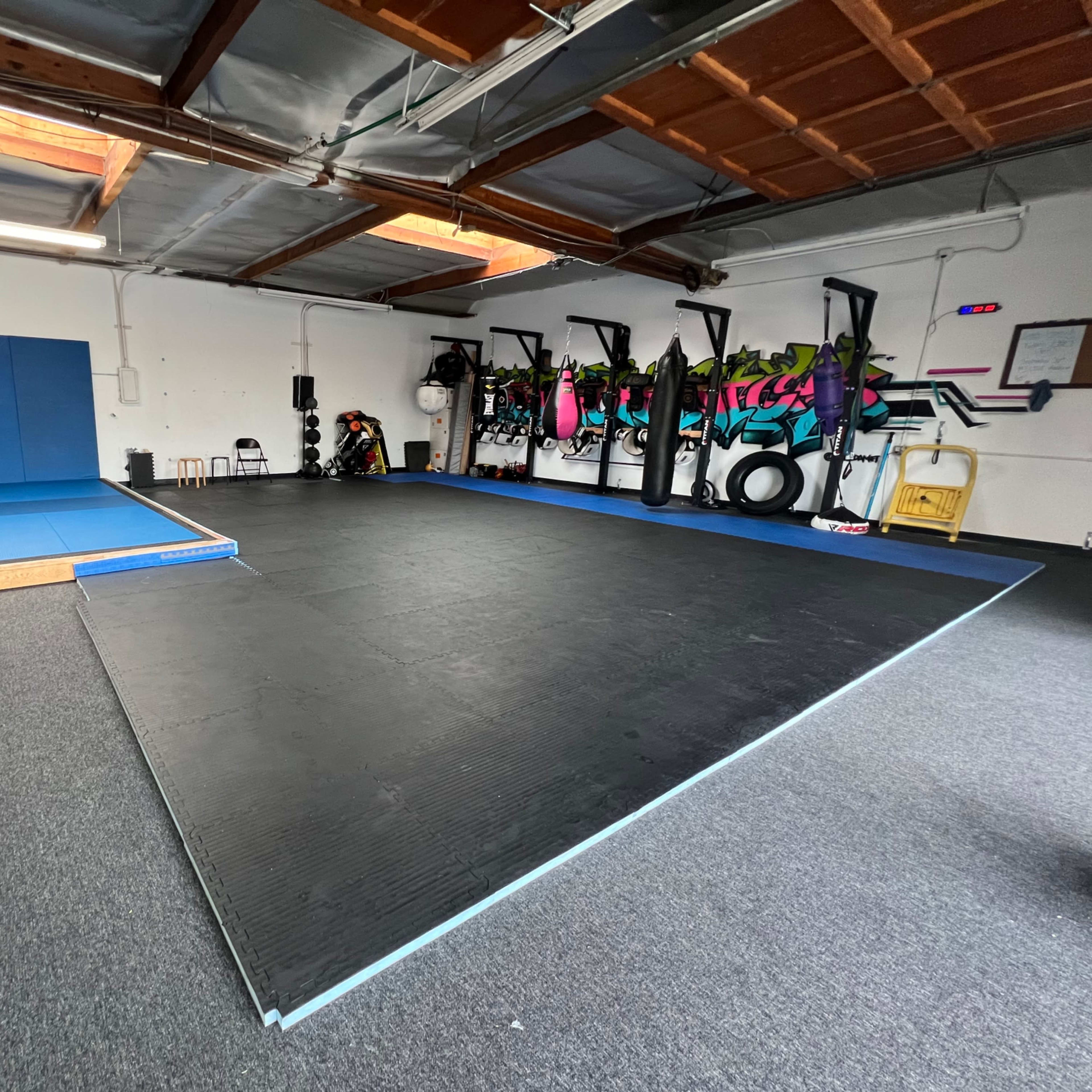 A training facility with a blue mat area and a black rubber floor, equipped with fitness equipment and wall decorations.