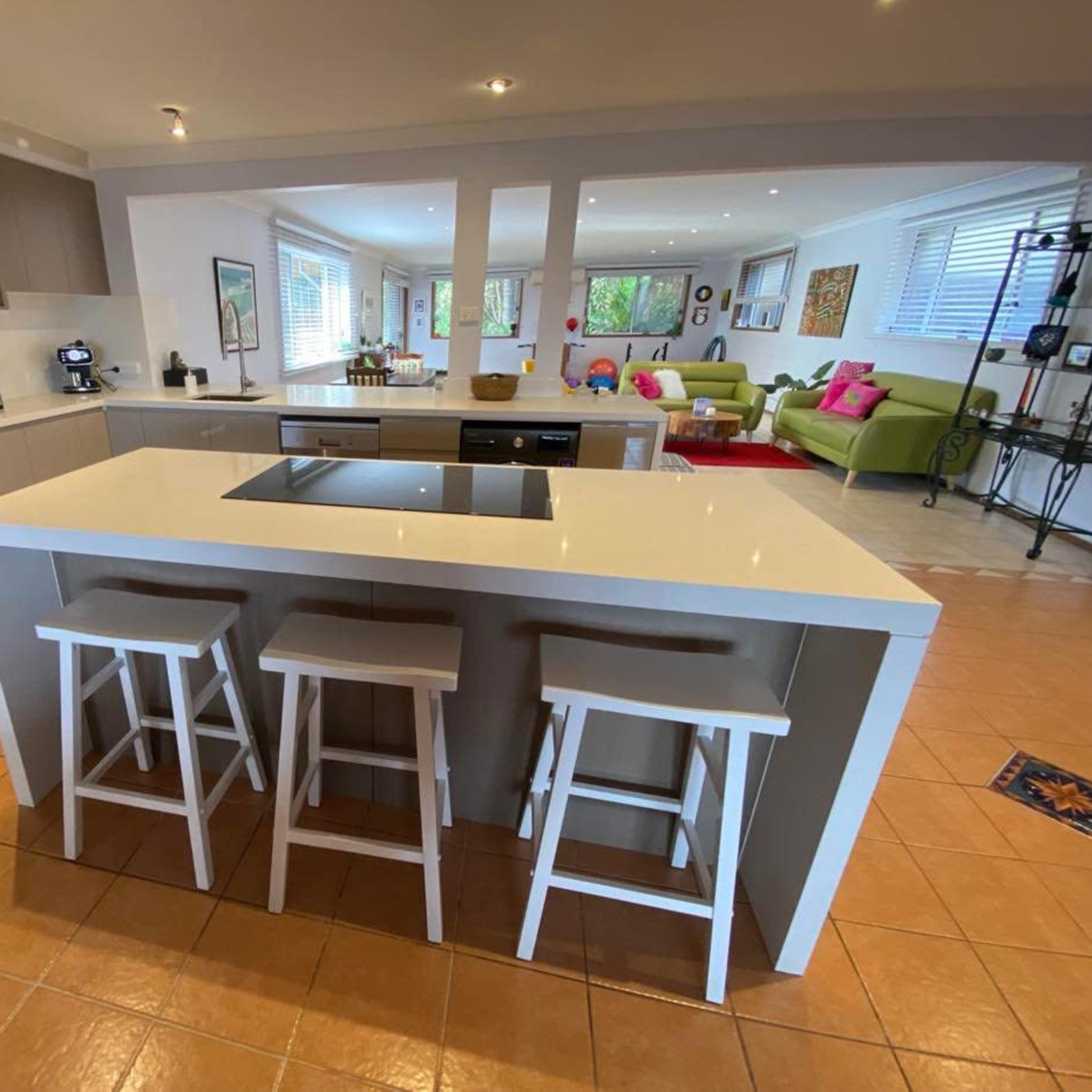 A modern kitchen with a central island, three white stools, stainless steel appliances, and an open layout leading to a living area.