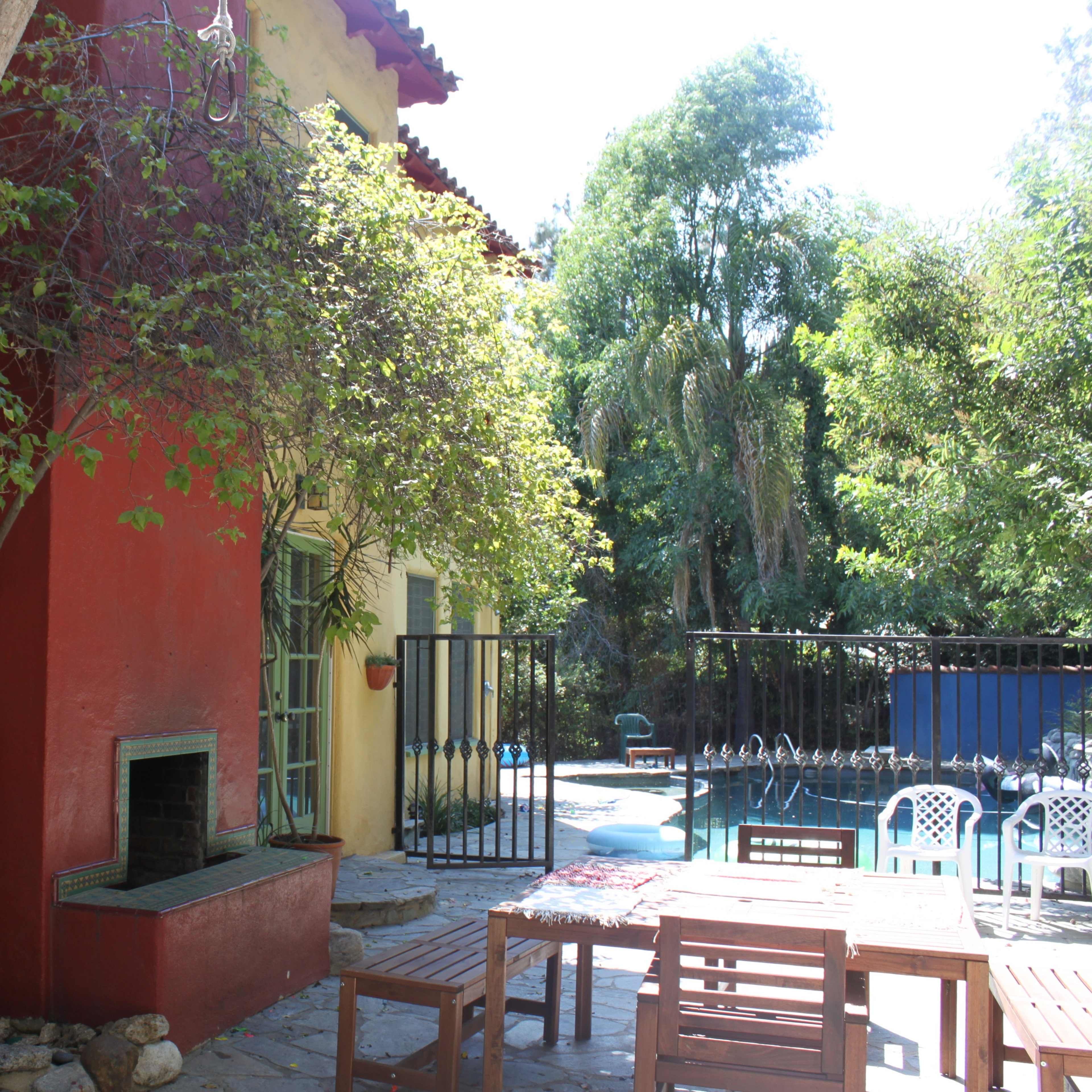 A terrace with wooden furniture is adjacent to a brightly colored house and a swimming pool surrounded by greenery.