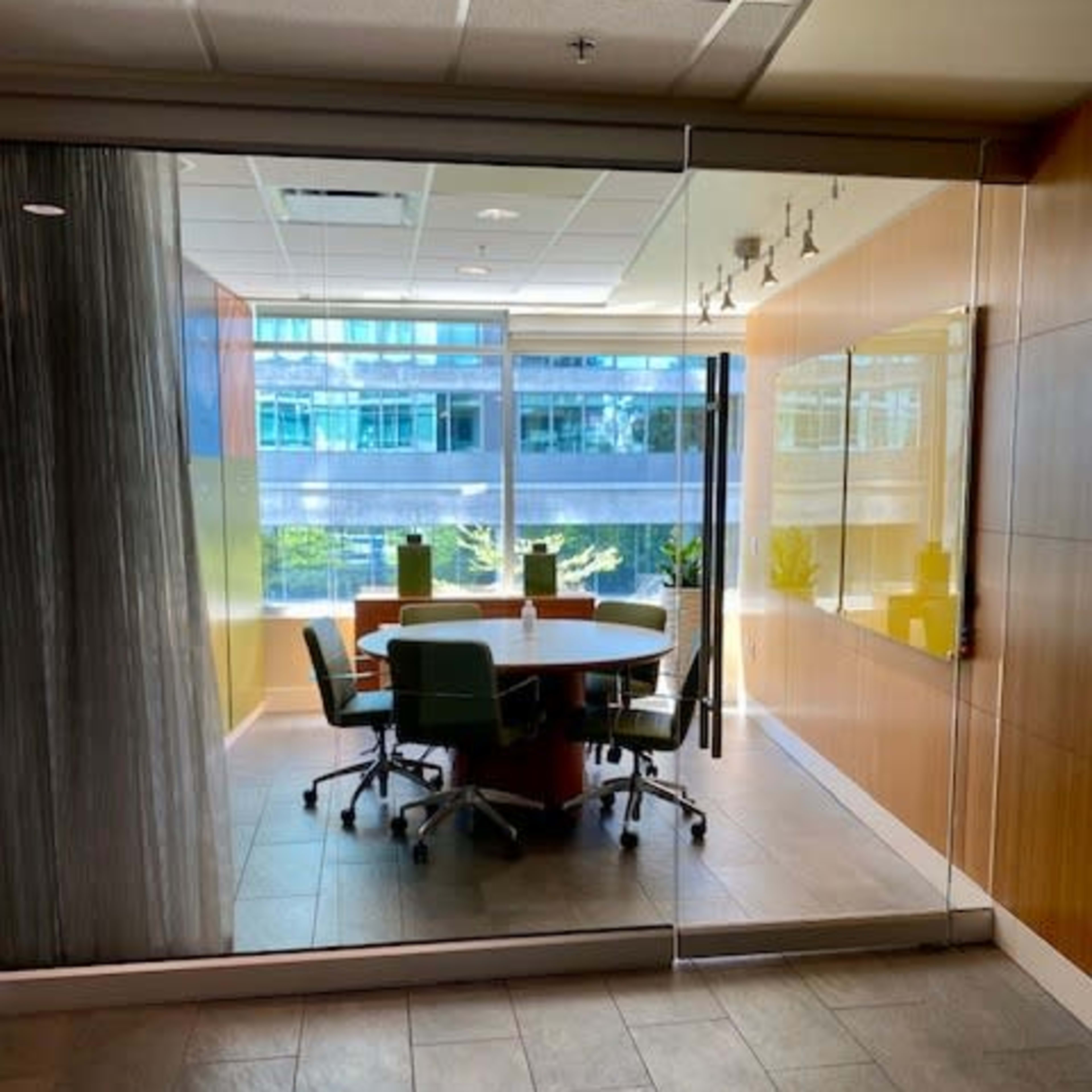 A glass-walled conference room features a round table surrounded by green chairs and large windows displaying the outside view.