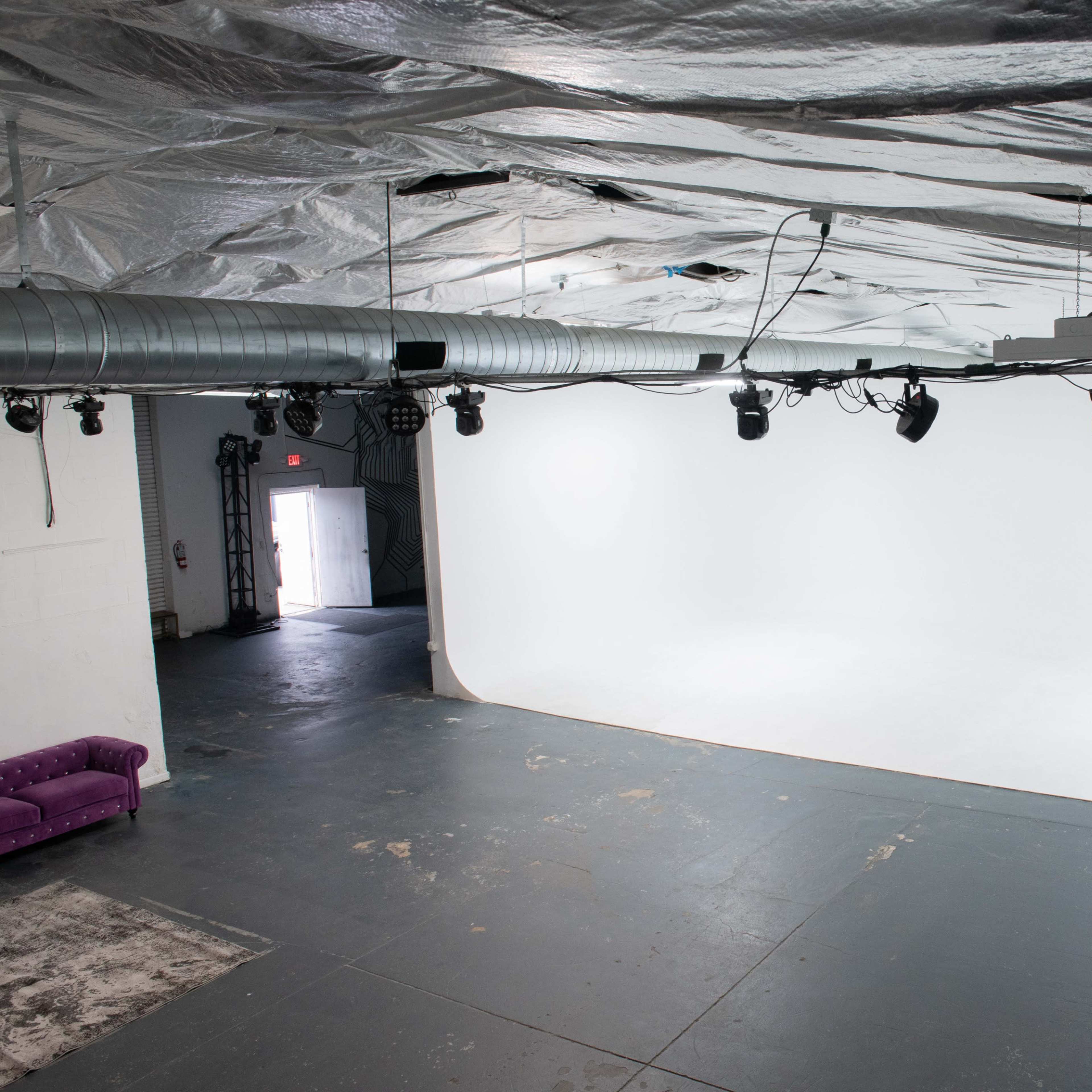 A large, empty studio space features a seamless white backdrop illuminated by ceiling-mounted lights, with a purple sofa in the corner.