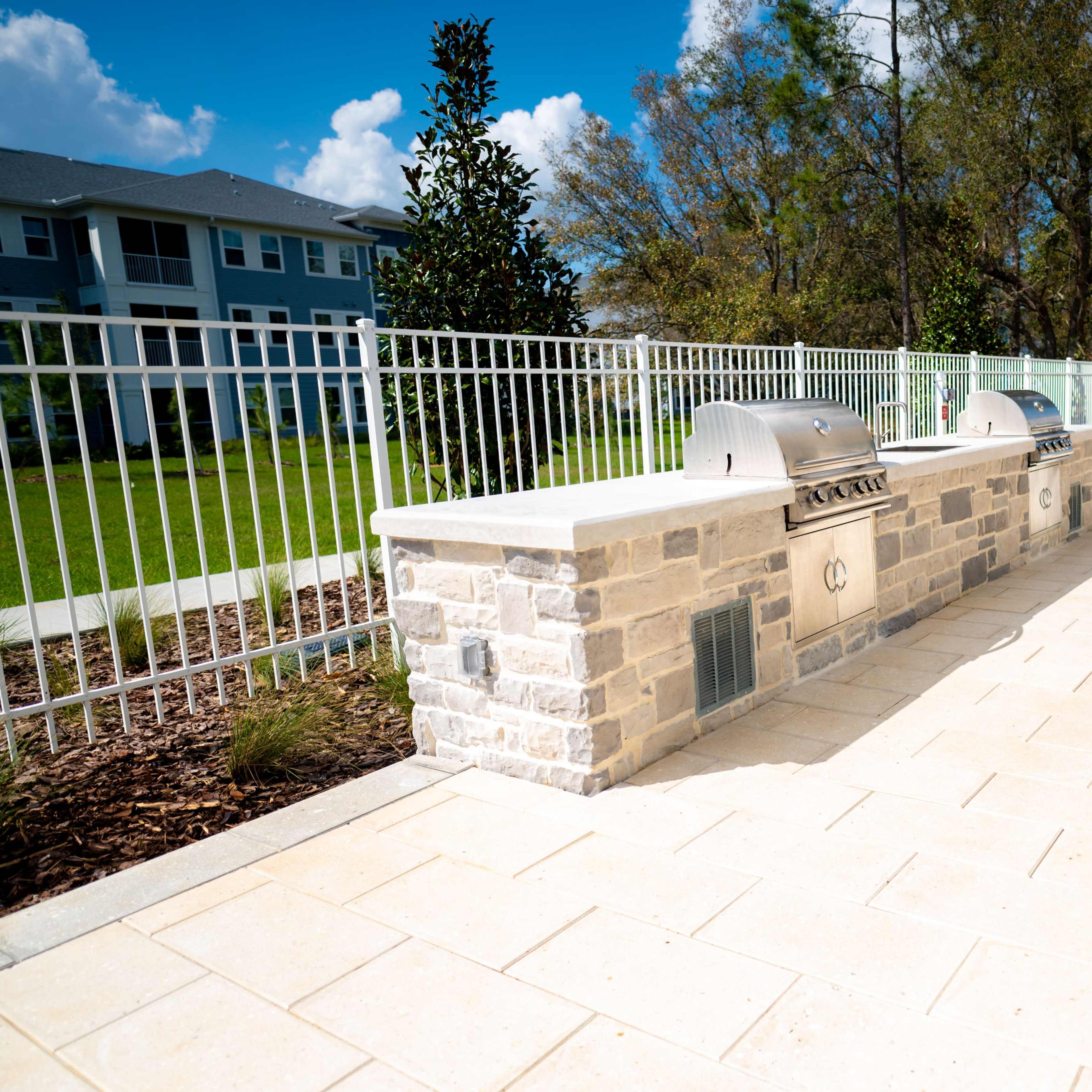 A stone outdoor grilling area with two metal grills, surrounded by a fence and landscaped green space.
