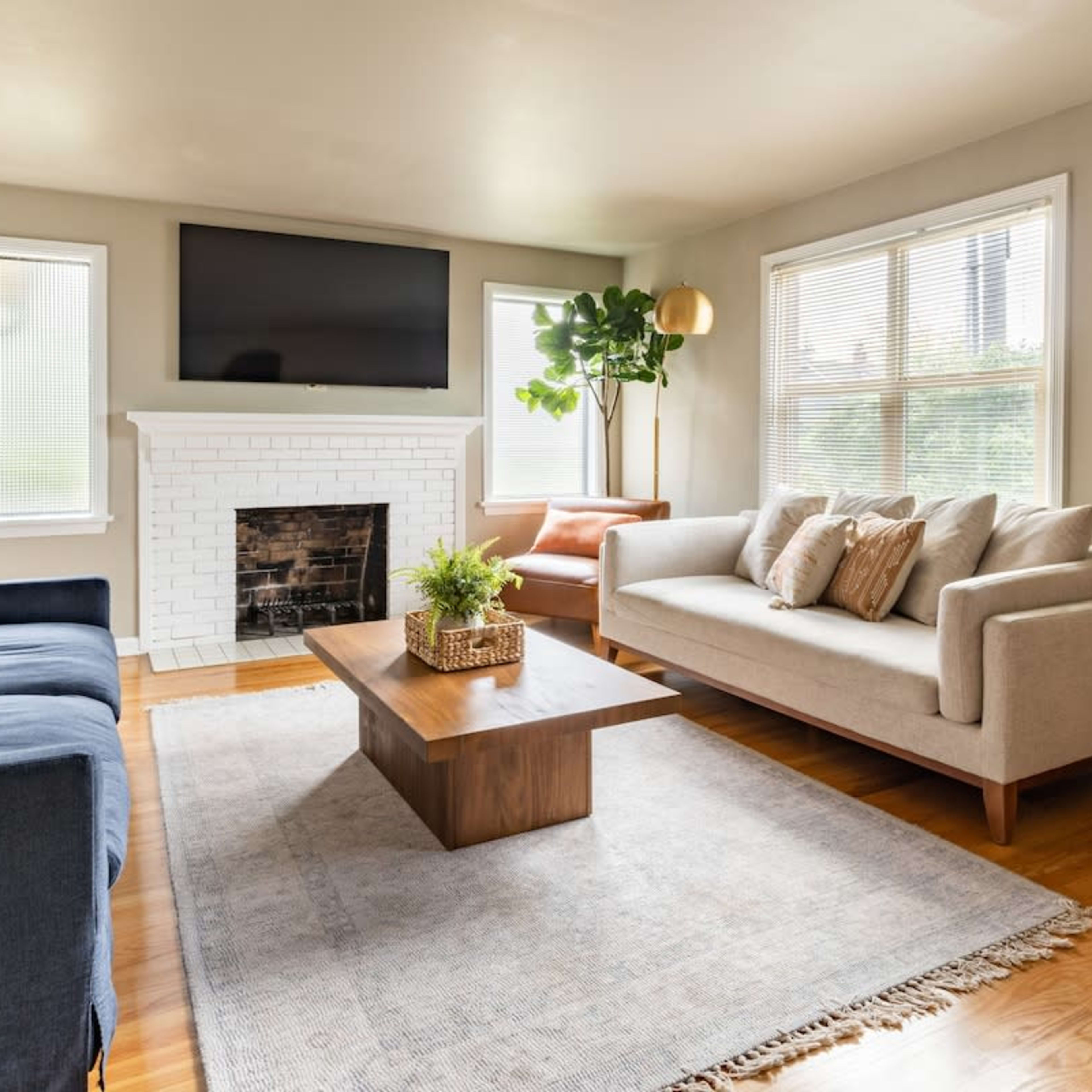 A living room features a blue couch, a gray sofa, a wooden coffee table, and a fireplace, with large windows allowing natural light to enter.