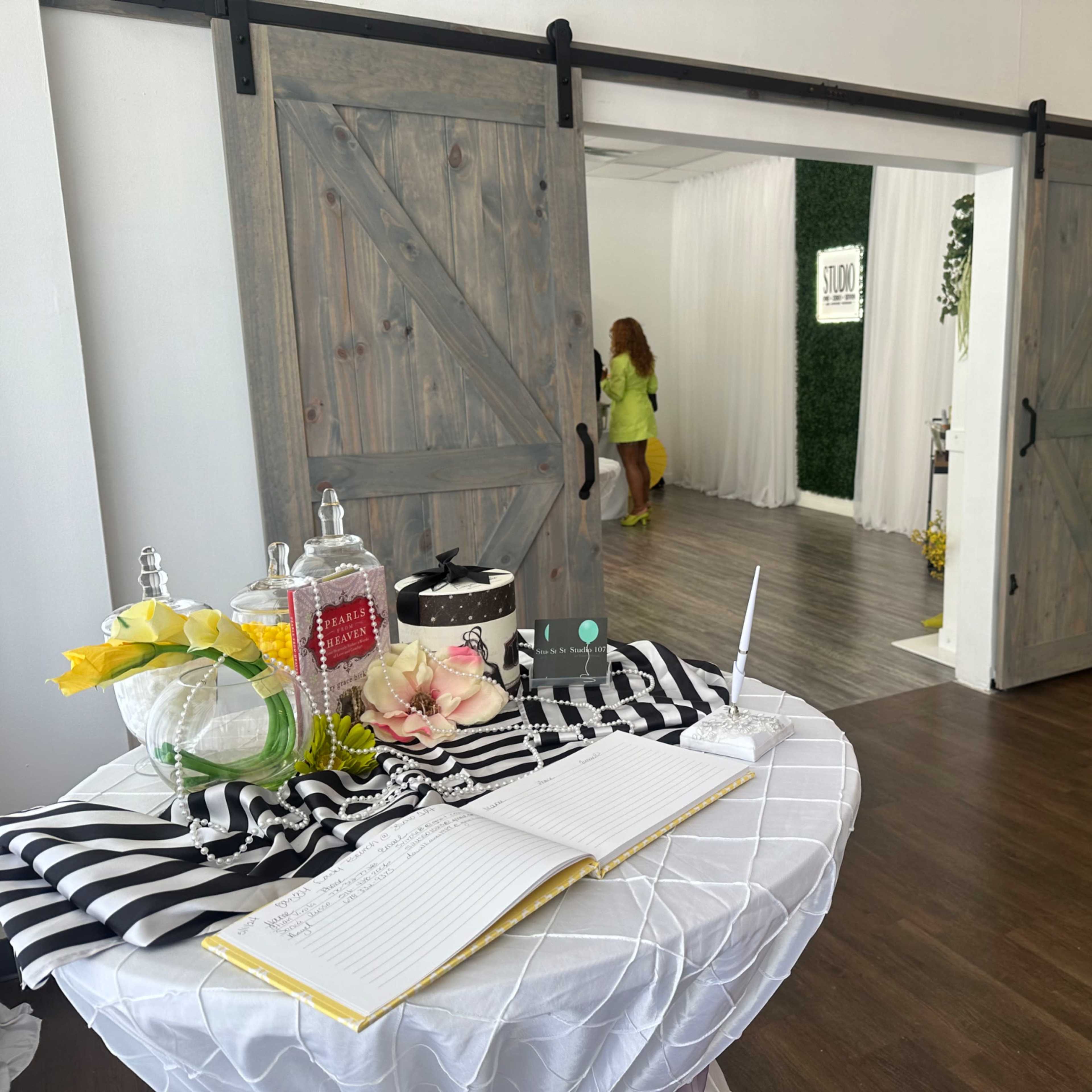 A bright event space features sliding barn doors, a decorated table with a guest book, and a backdrop adorned with colorful balloons.