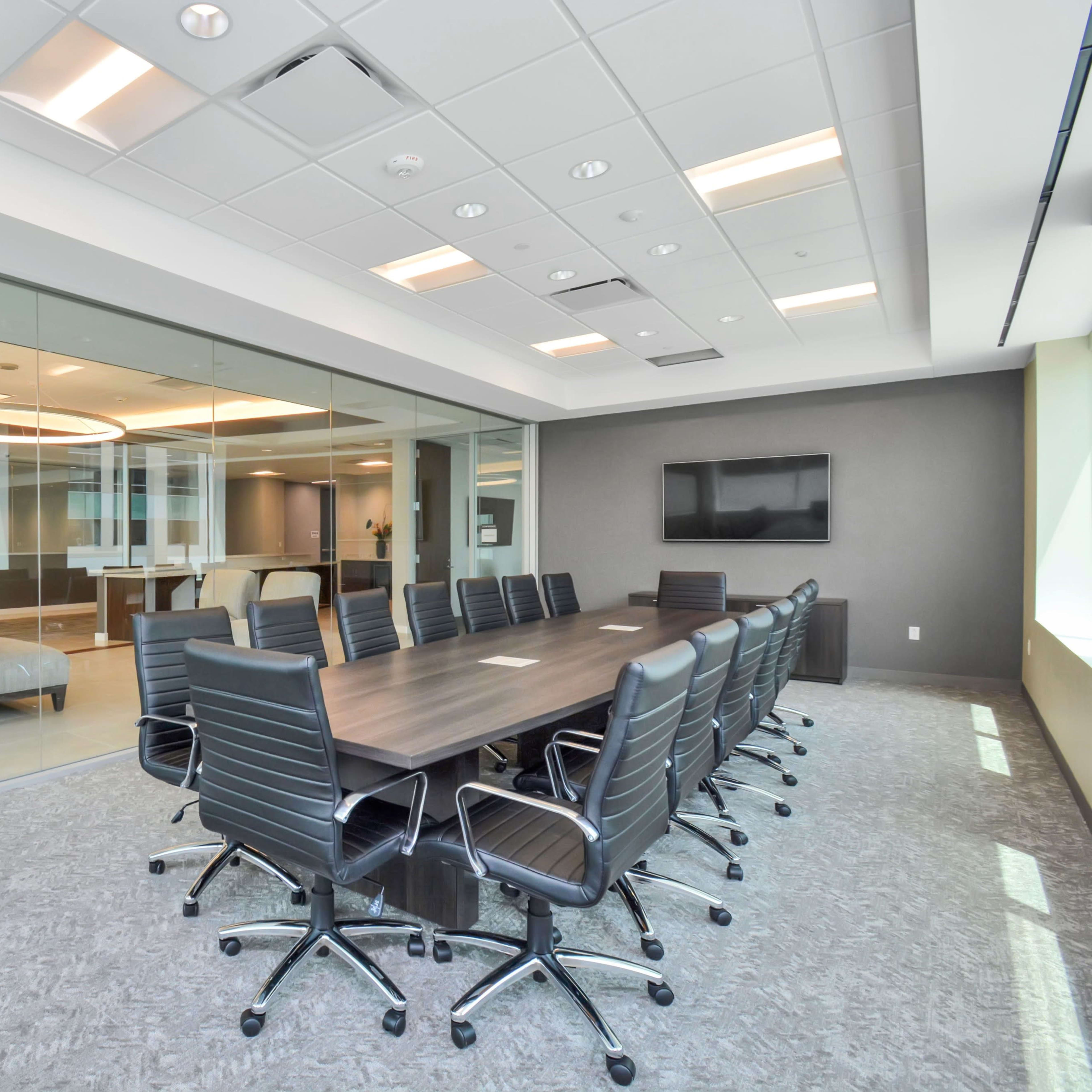 A modern conference room with a large wooden table surrounded by black office chairs and a glass wall leading to another meeting area.