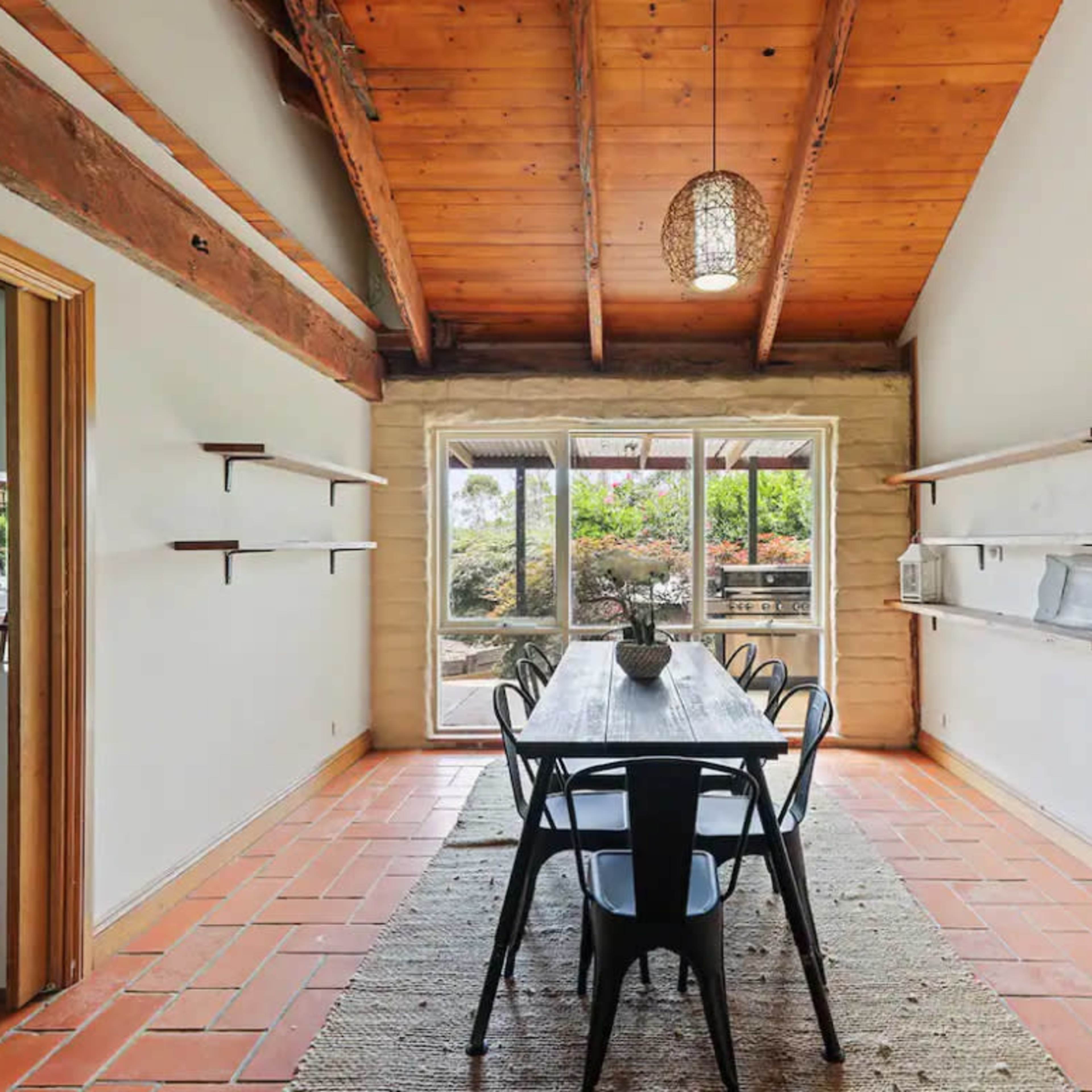A long dining table with black chairs sits in a bright room featuring exposed wooden beams and large windows that overlook a garden.