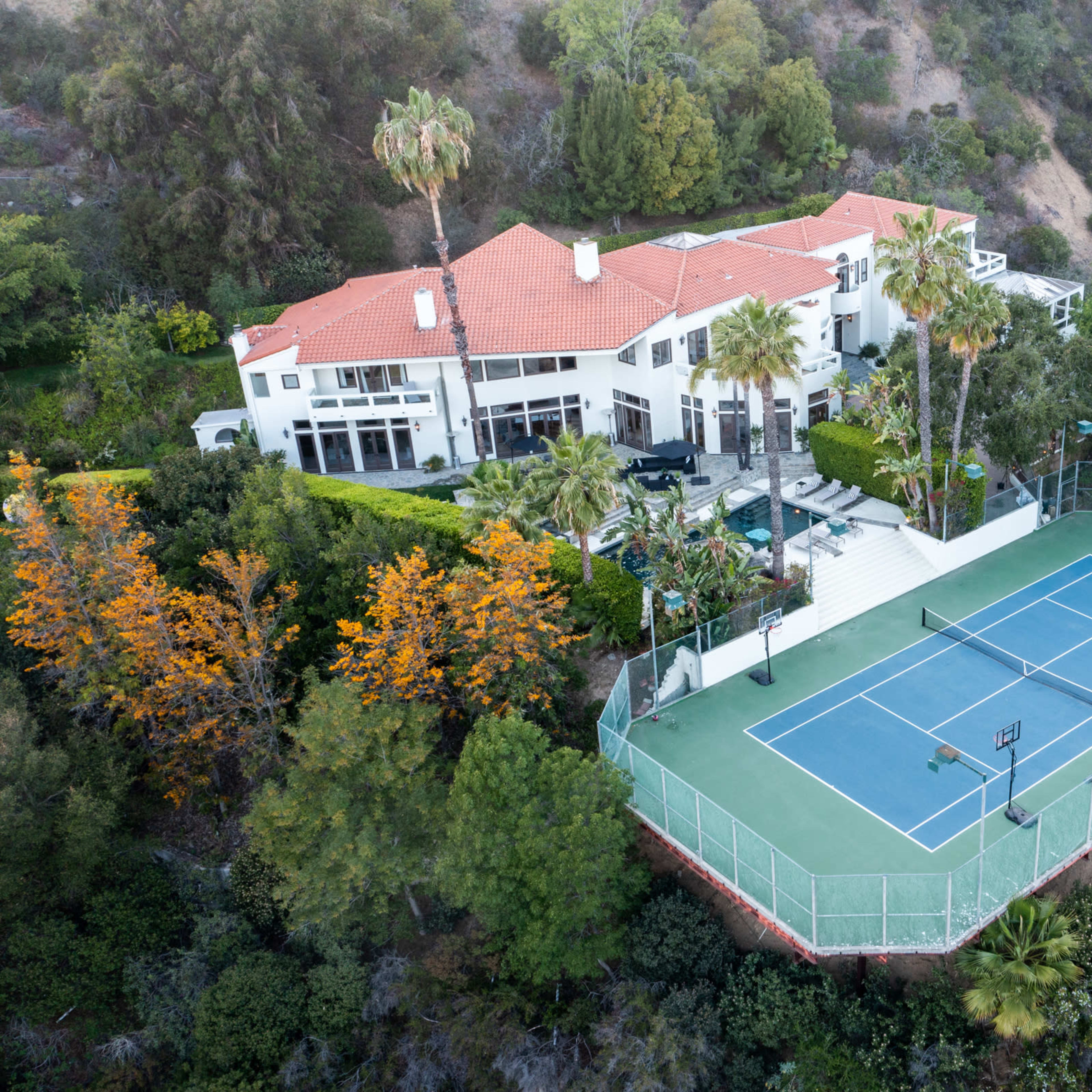 A large house with a red-tiled roof surrounded by greenery and a tennis court nearby.