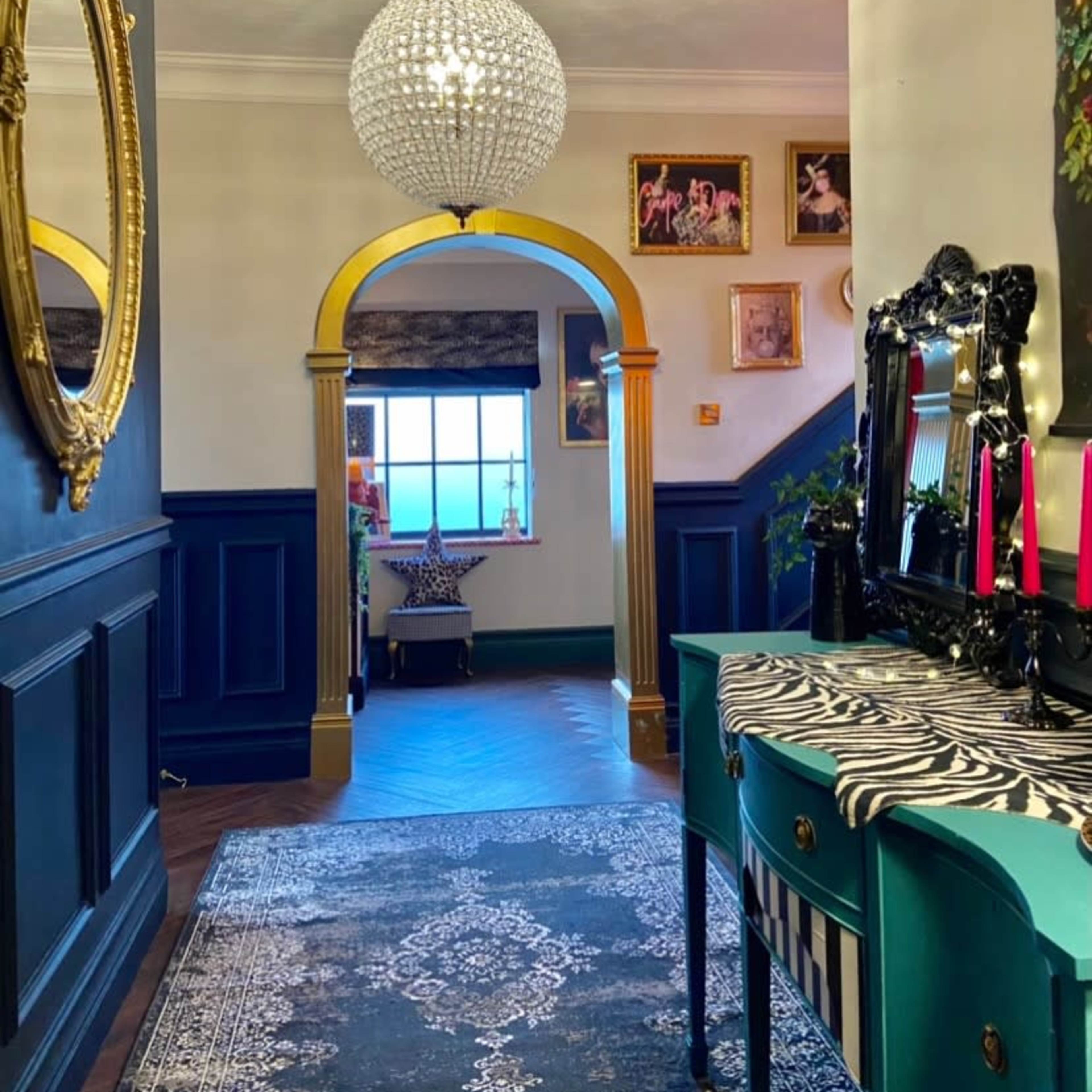 A hallway with dark blue walls, a round chandelier, and an ornate mirror, leading to an archway and a staircase.