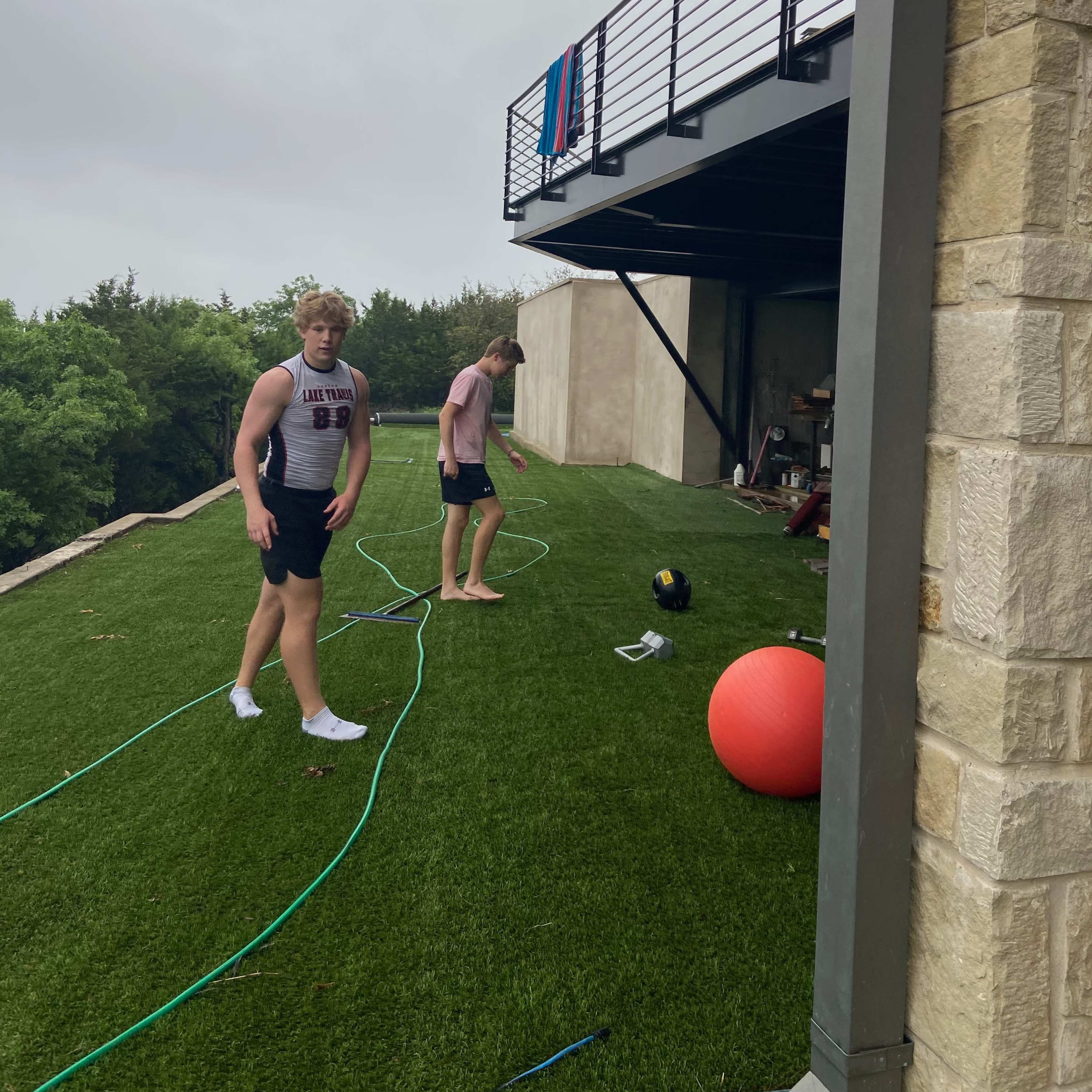 Two young men stand on a green lawn next to a building with a balcony, one of them holding a hose while the other is near a large red ball.