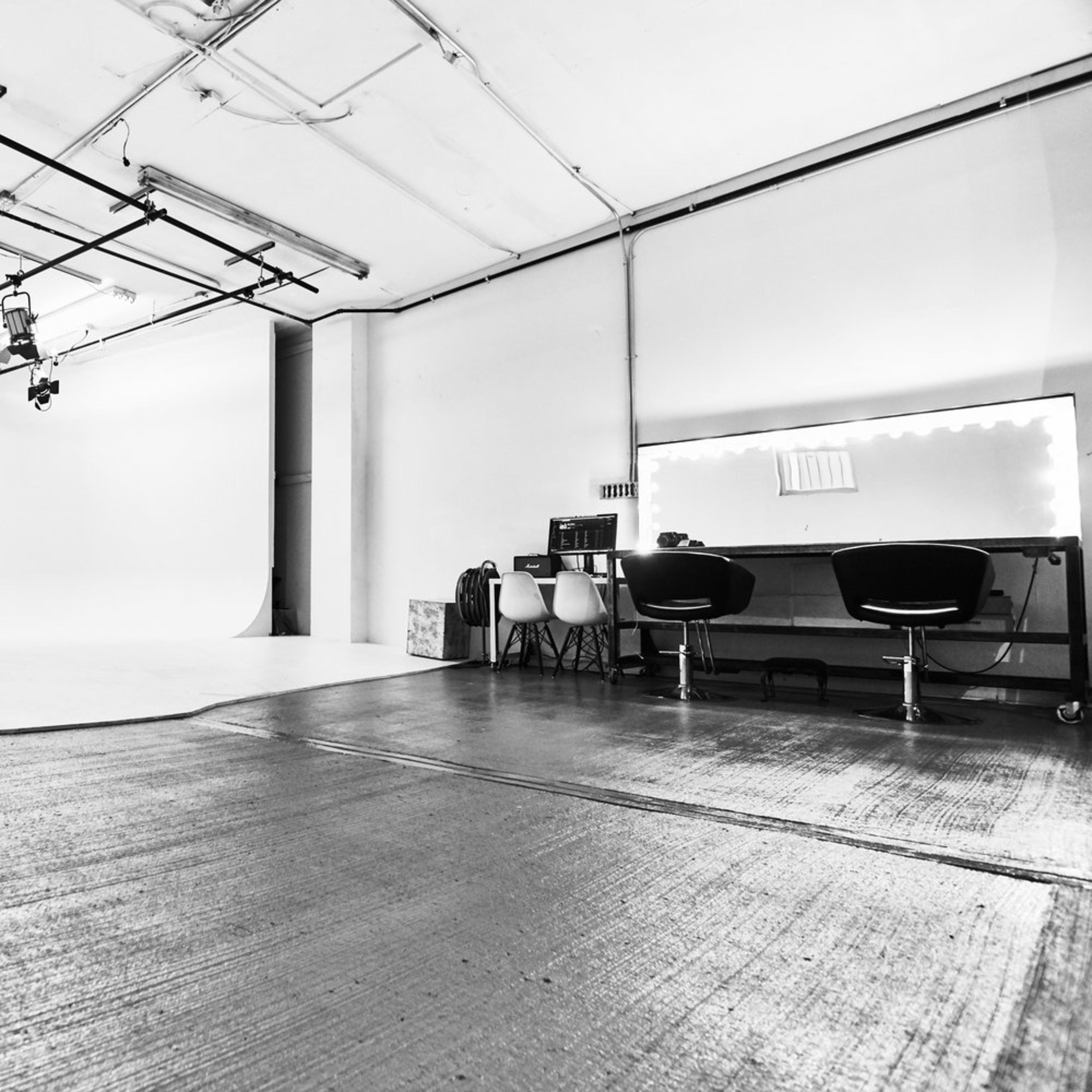 A minimalist photography studio with a white backdrop, overhead lighting, and a row of chairs beside a vanity mirror.