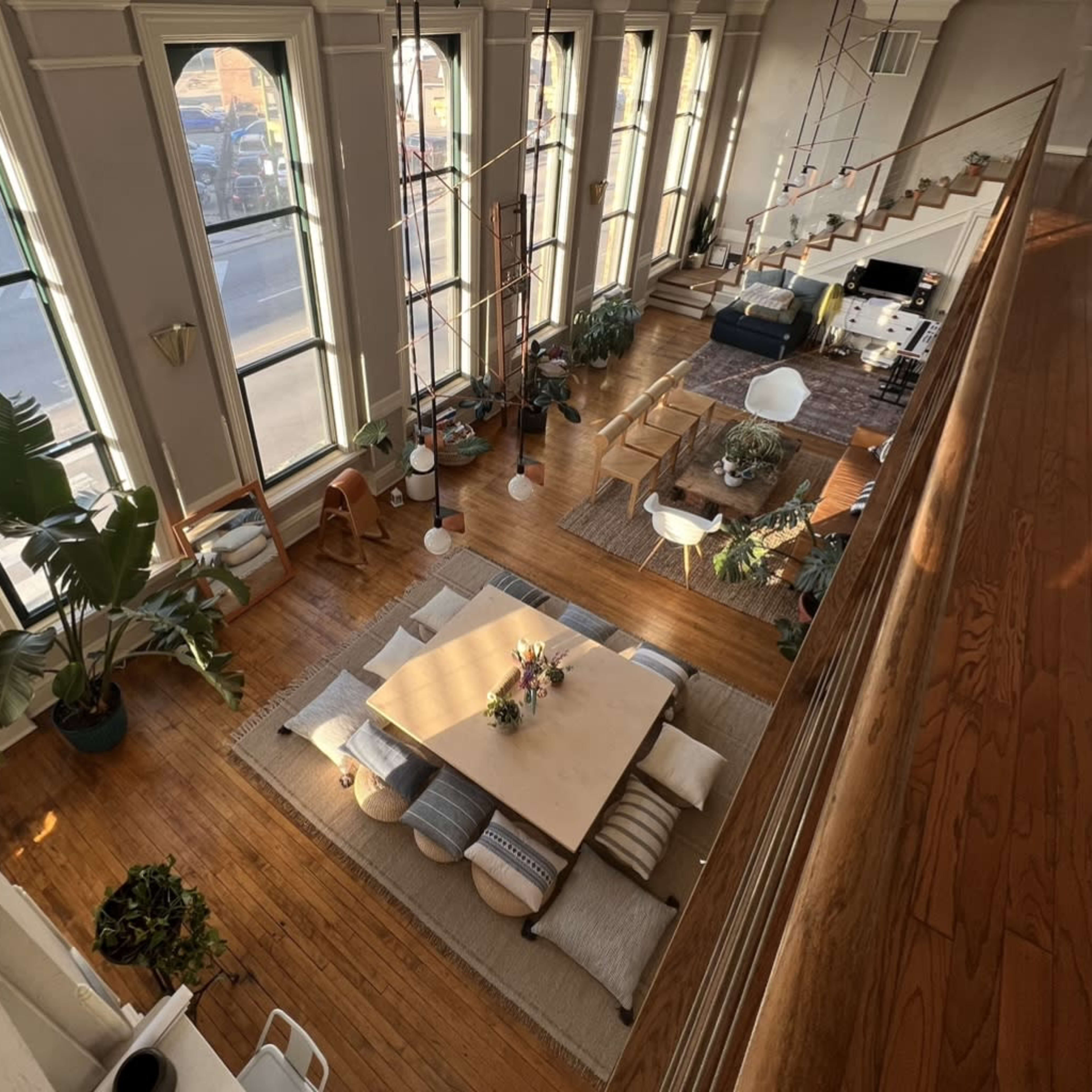 A spacious, well-lit living area with large windows, wooden flooring, a central low table surrounded by cushions, and various plants placed throughout.