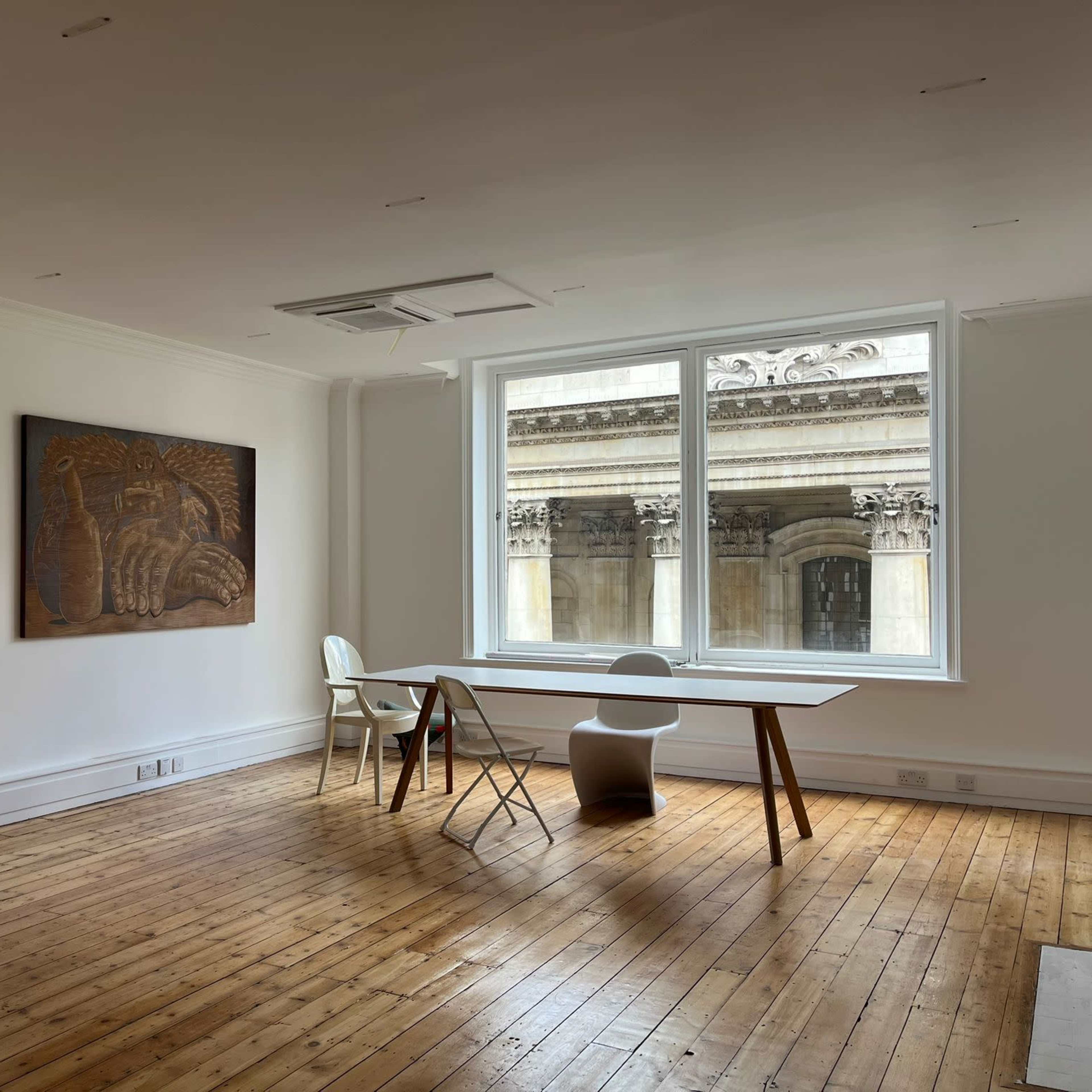 A spacious, empty room with wooden floors, a large window featuring a view of an exterior architectural detail, a wooden table, and a white chair.