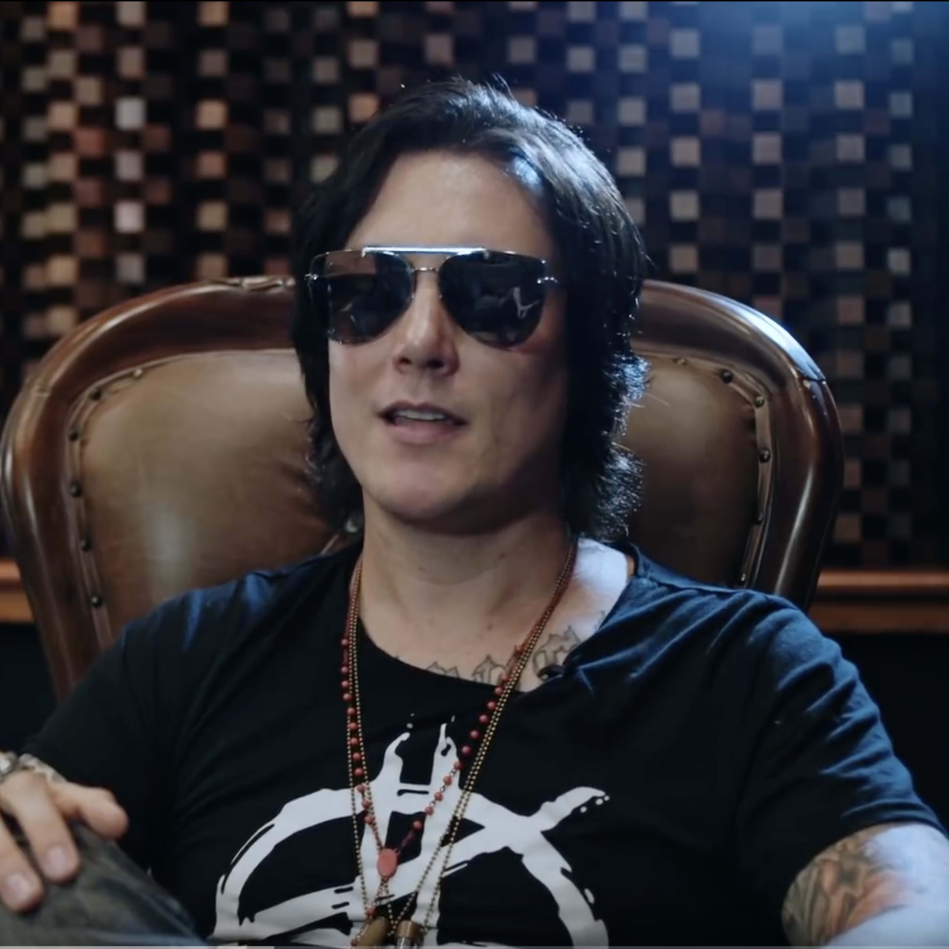 A musician wearing sunglasses sits in a chair in a studio with a patterned wall and recording equipment in the background.