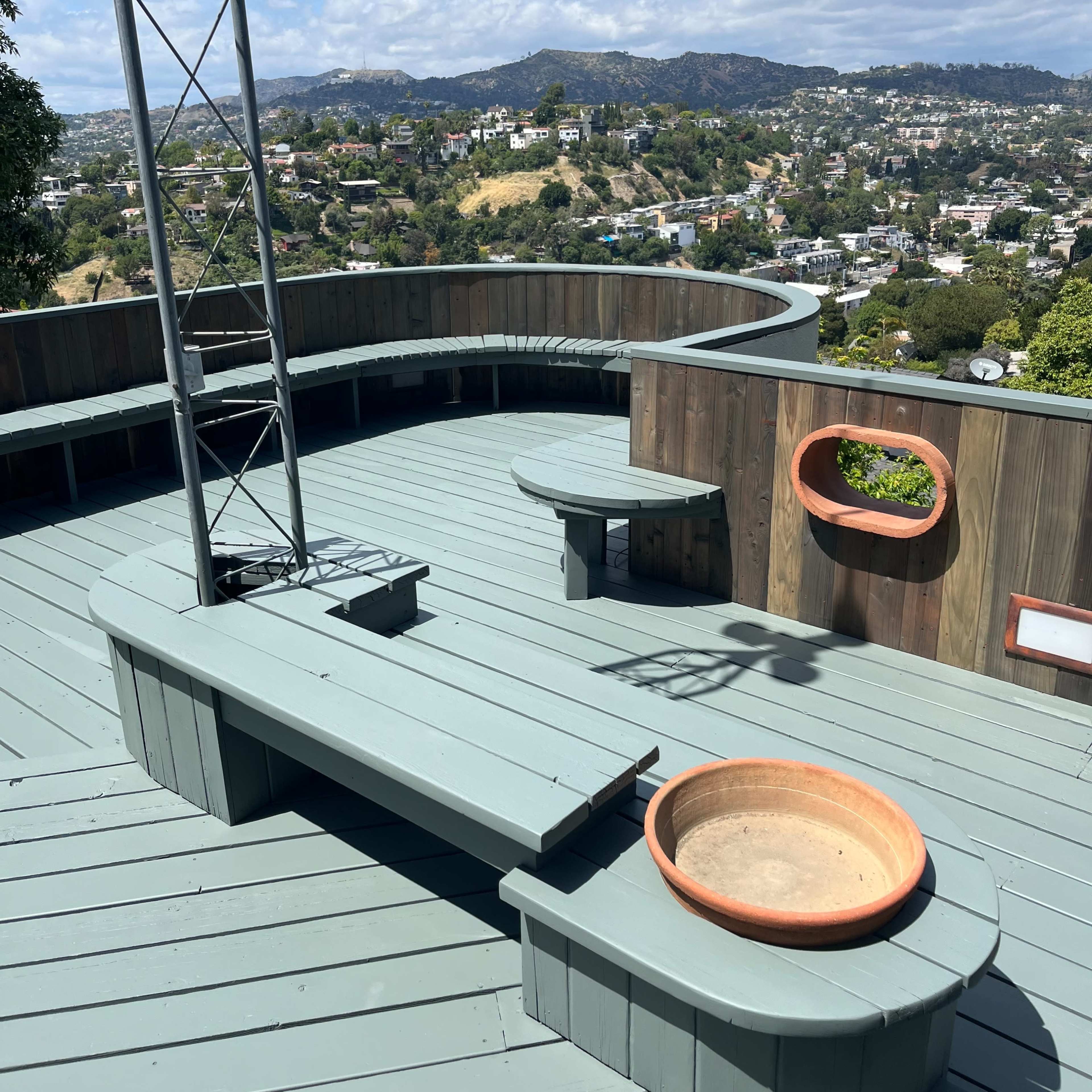 A wooden deck with a curved seating area, a fire pit, and a view of hills and buildings in the background.
